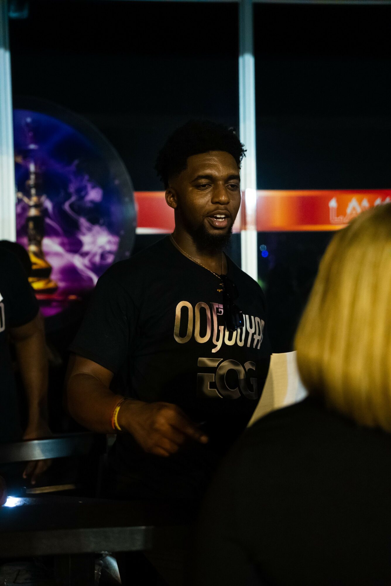 Young man with dark skin and short hair speaking at an event, with a colorful background and a person with blonde hair in foreground.
