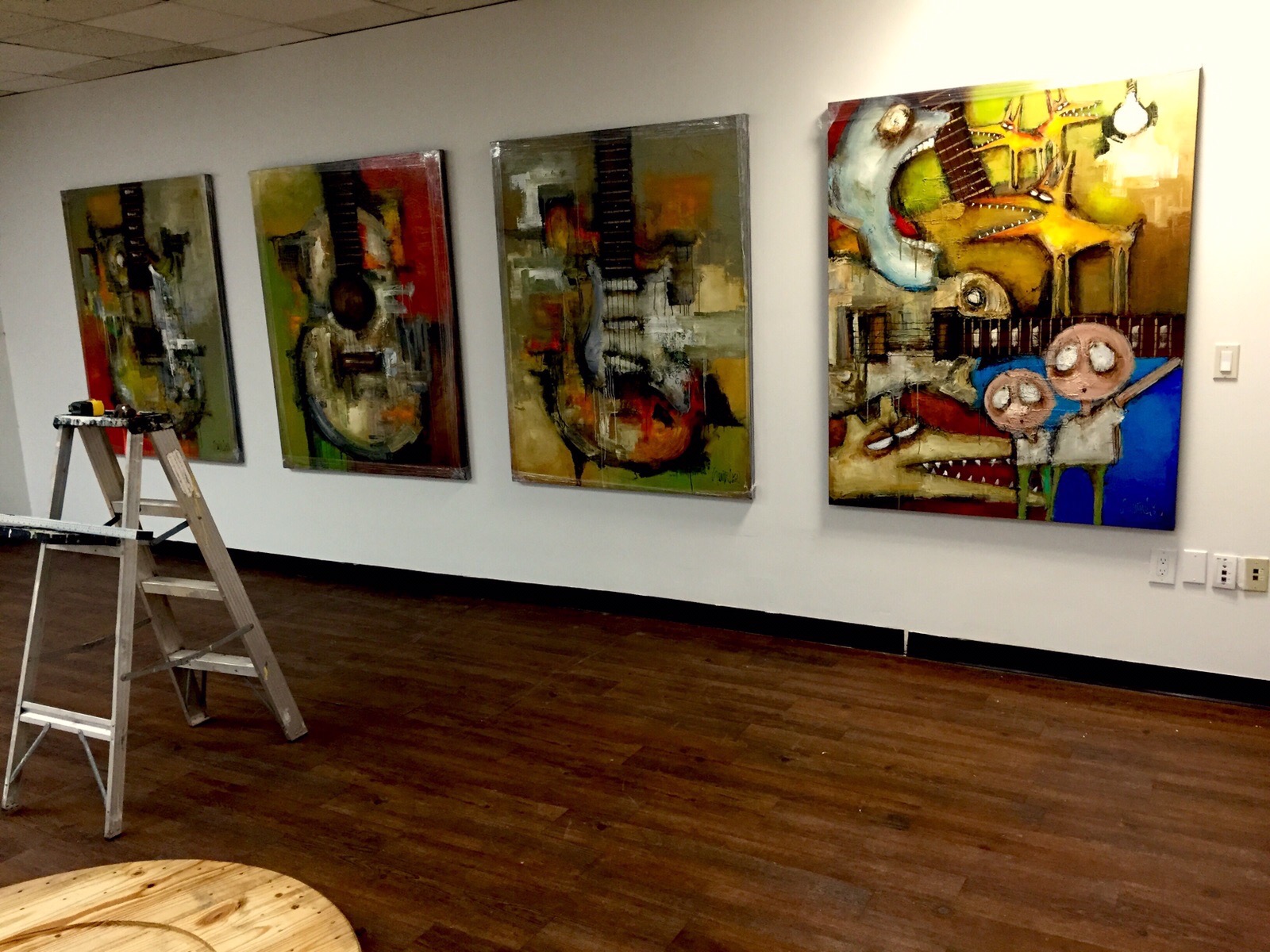 Four abstract paintings on a white gallery wall, with a ladder and wooden floor visible.