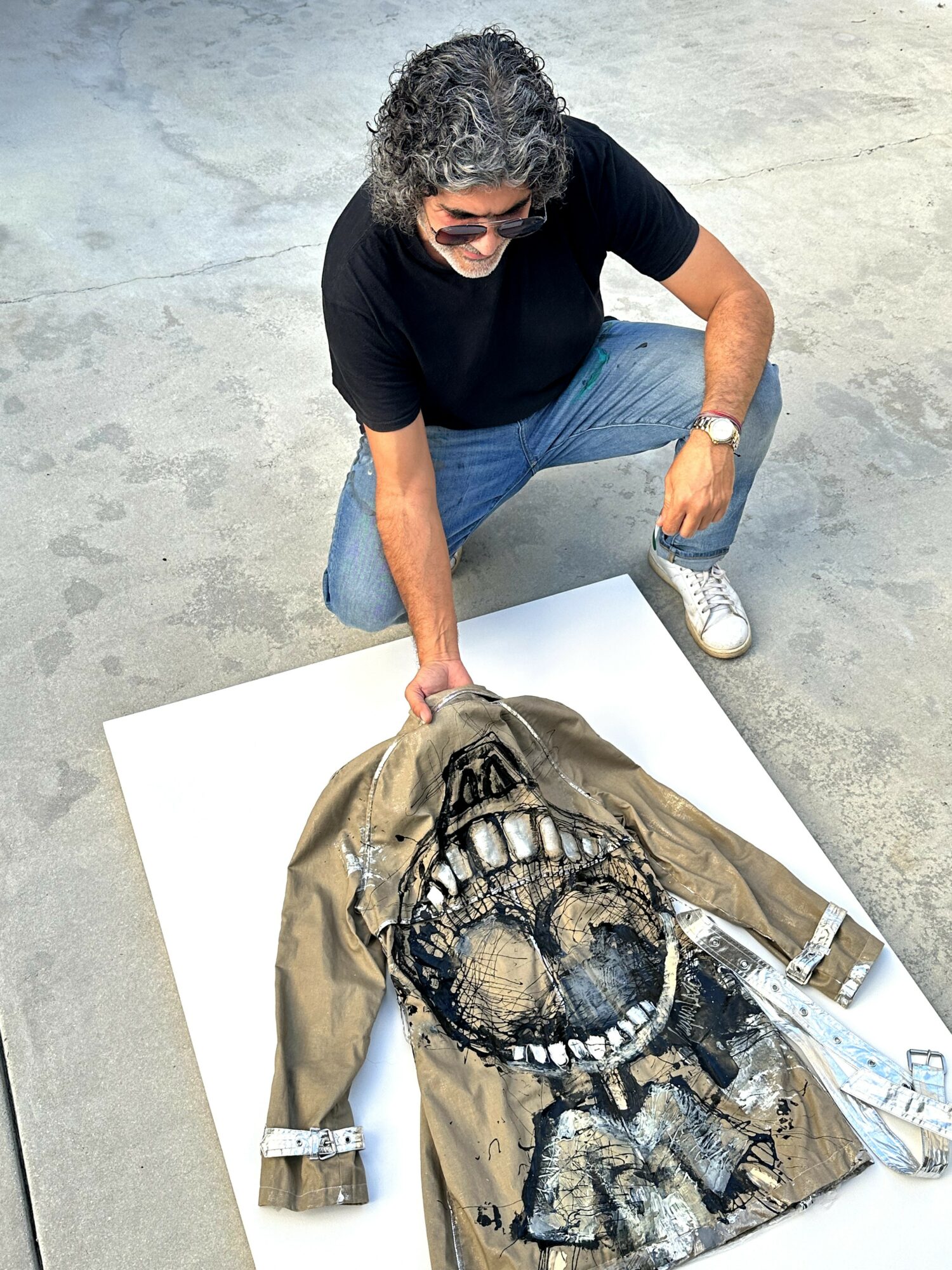 Person with curly hair and sunglasses crouches next to a large, detailed dinosaur costume on a white surface.