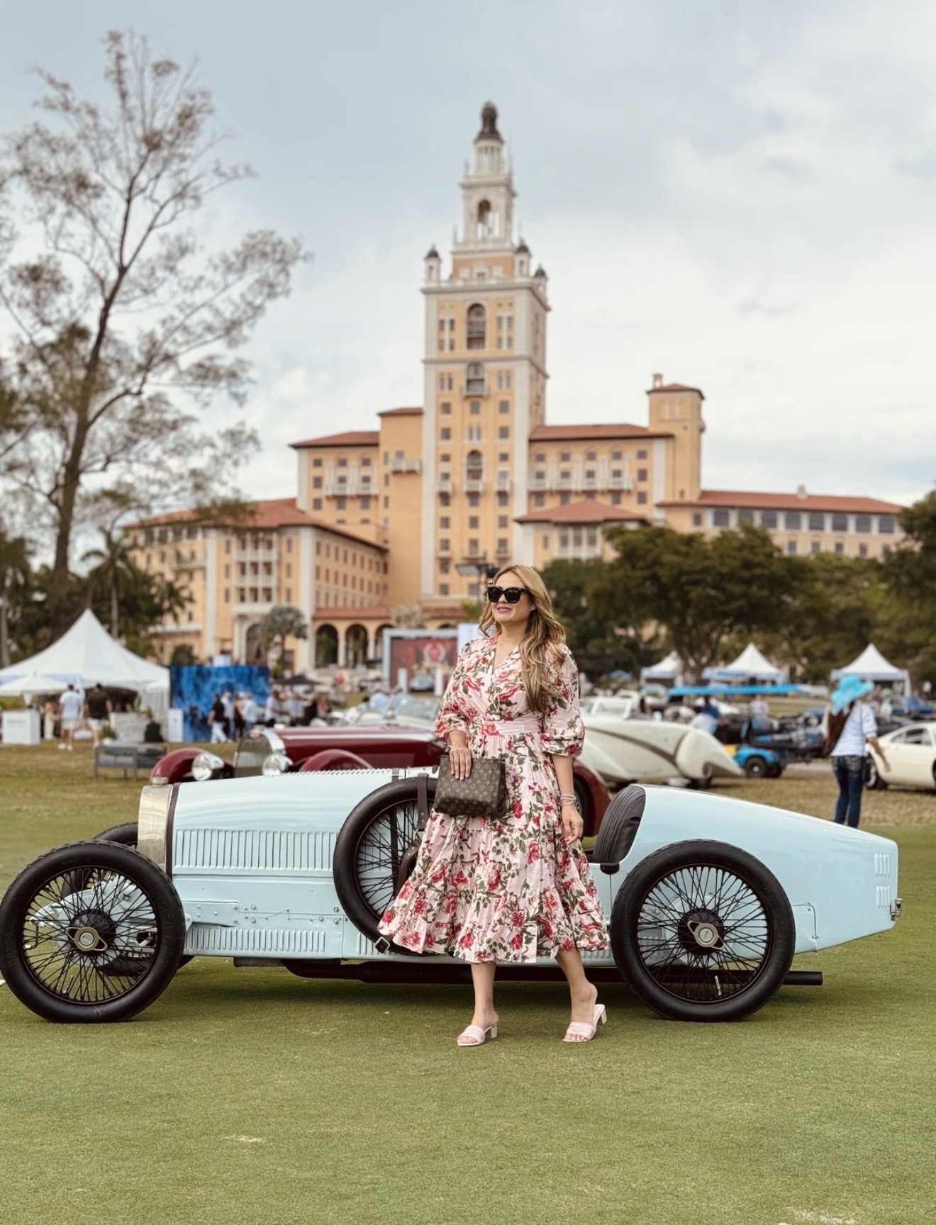 Woman in floral dress and sunglasses standing next to vintage car outdoors with a large building in the background.