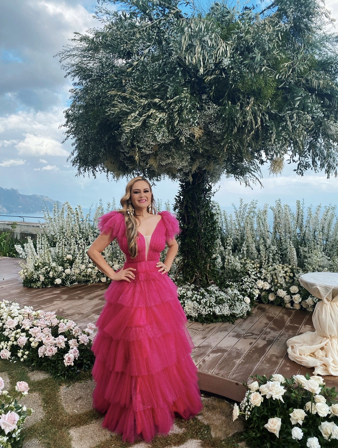 Woman in a bright pink gown standing outdoors near a large tree and flower arrangements, with a body of water in the background.