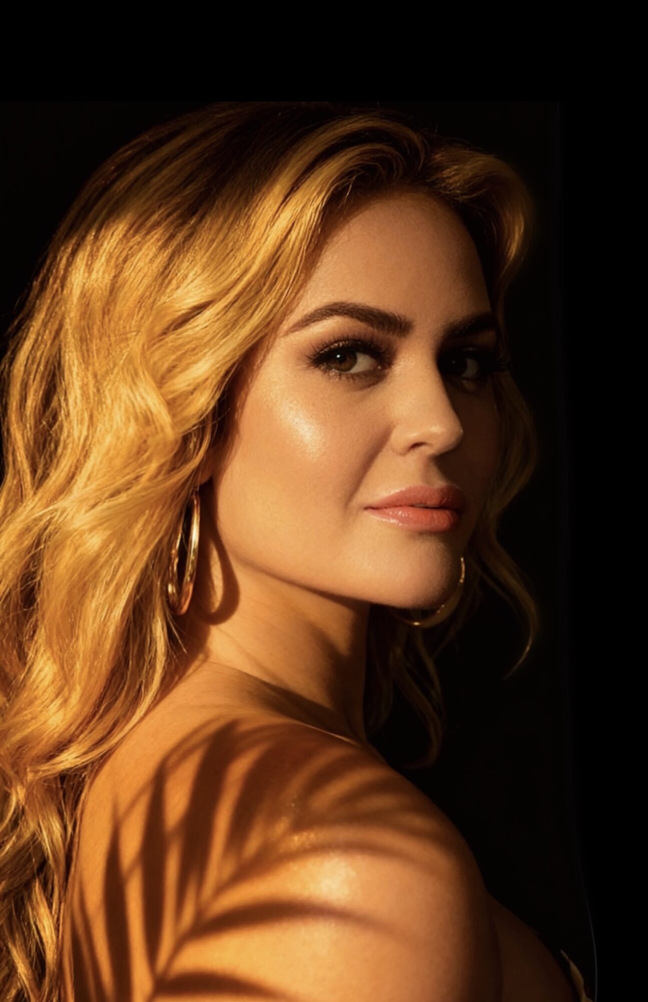 Portrait of a woman with wavy blonde hair, looking sideways, with shadow patterns on her shoulder, against a dark background.