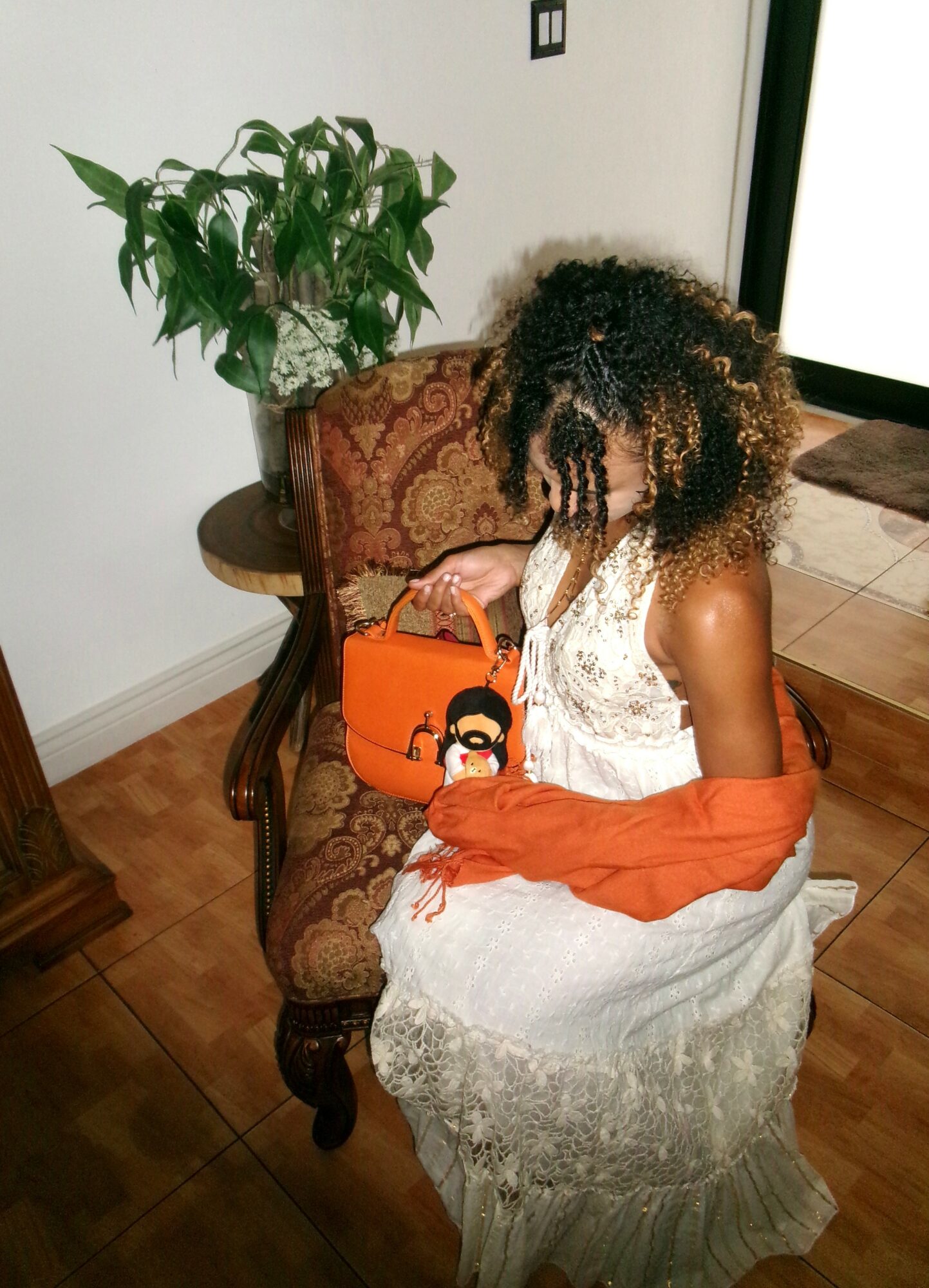 Woman with curly hair sitting on a patterned chair, holding an orange bag, in a room with a plant and wooden furniture.