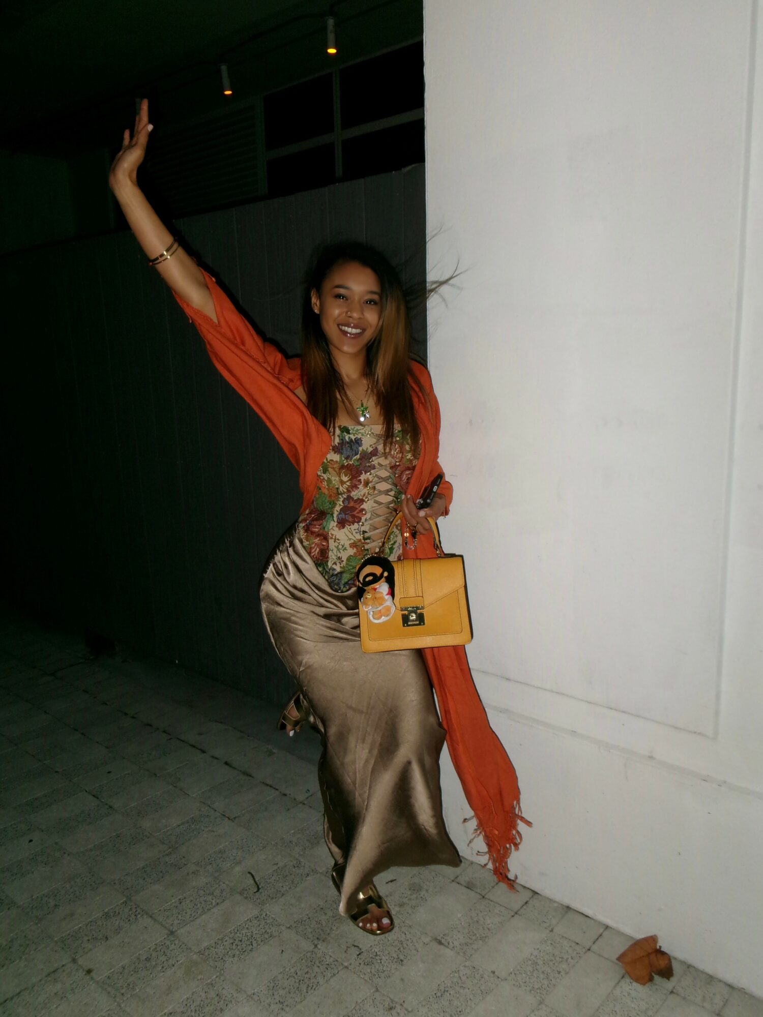 Woman in colorful dress and orange shawl smiling, holding a yellow purse, standing against a white wall.