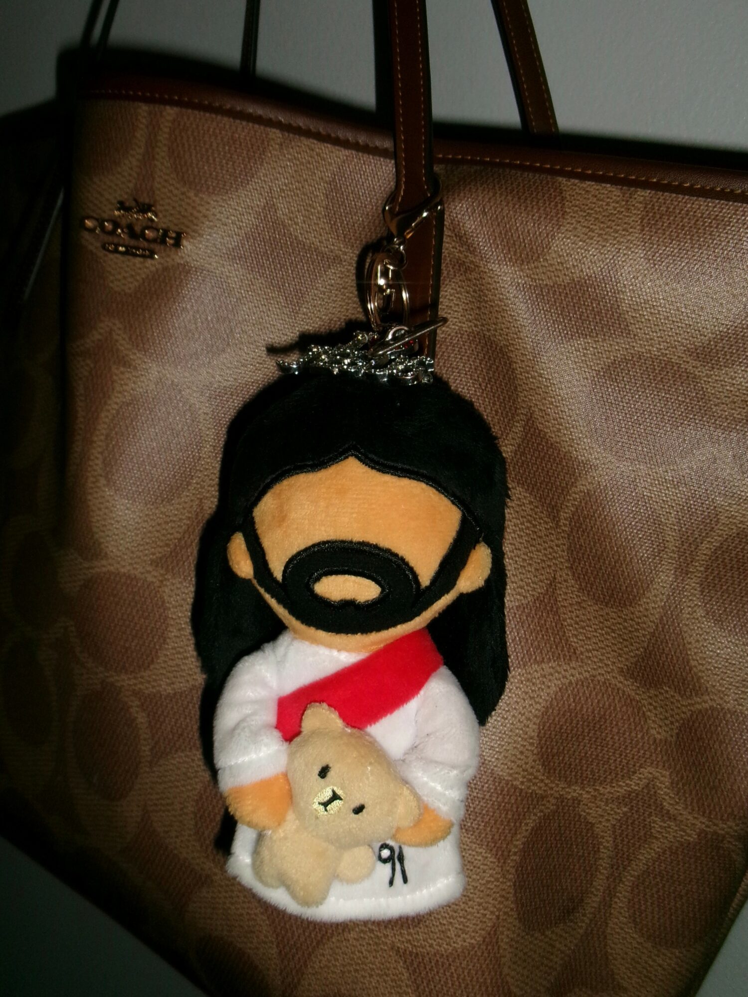 Keychain plush toy of a person with long black hair, beard, white robe, red sash, holding a small beige animal, hanging from a bag