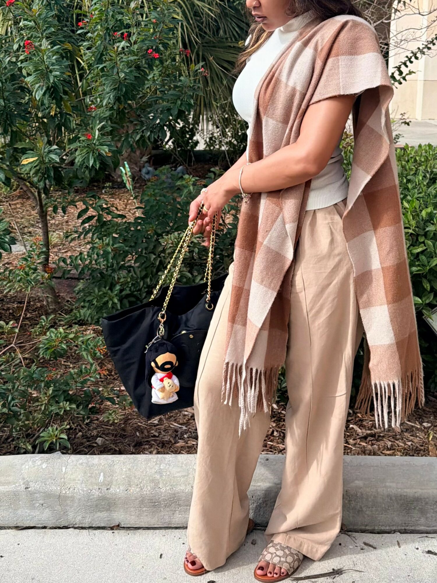 Woman standing outdoors with greenery, wearing beige pants and a large checkered scarf, holding a black bag with a plush toy keychain.