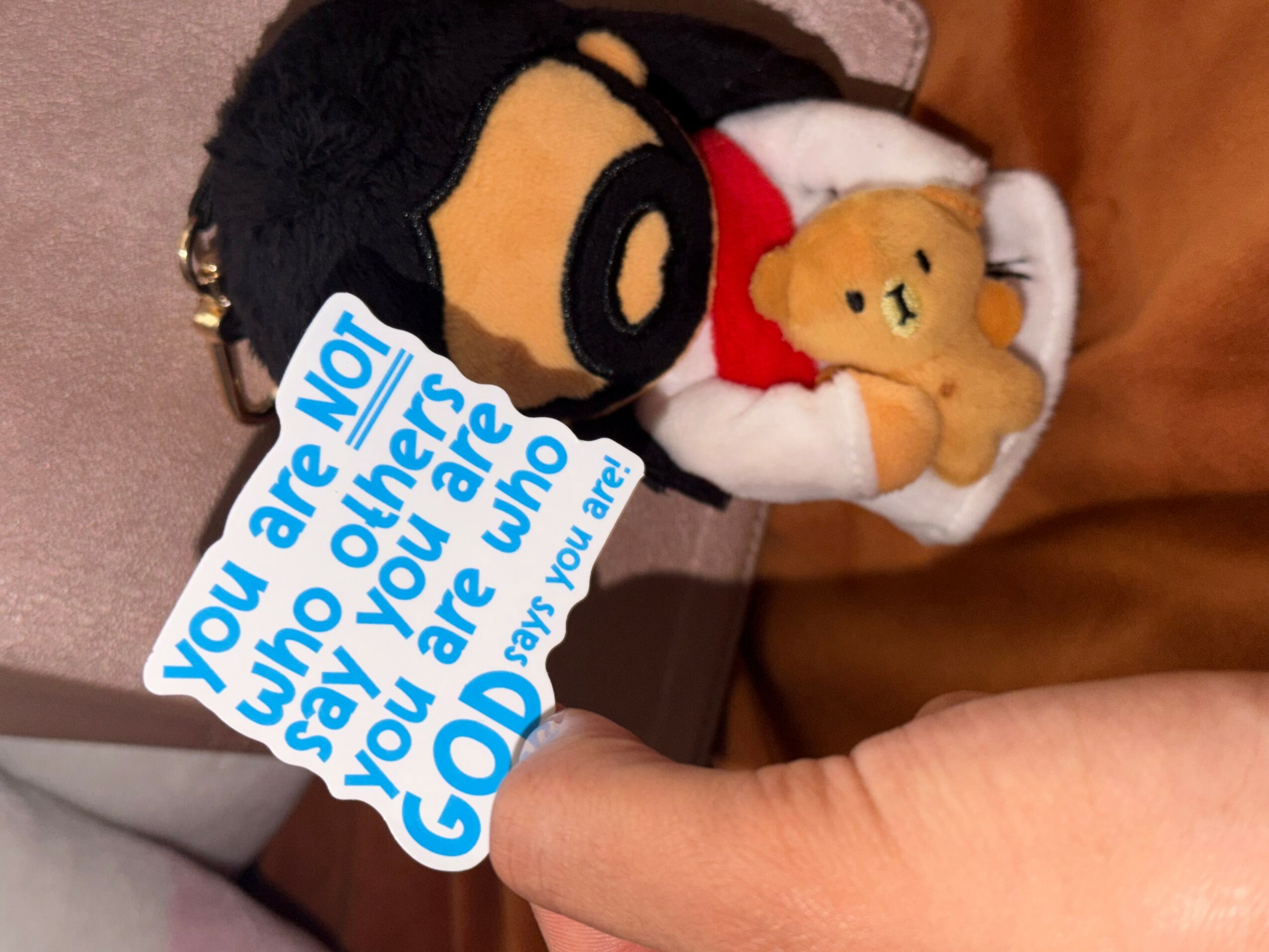 Person holding a plush teddy bear and a small stuffed animal, with a blue and white note partially covering the plush toy.