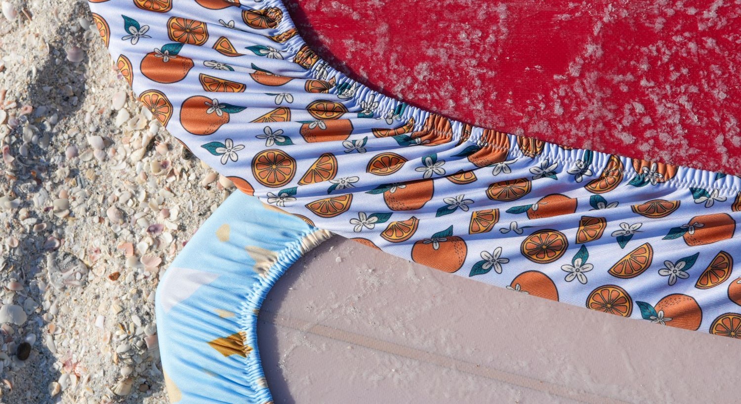 Close-up of a beach towel with a red section, patterned fabric, and sand, on a sandy surface.