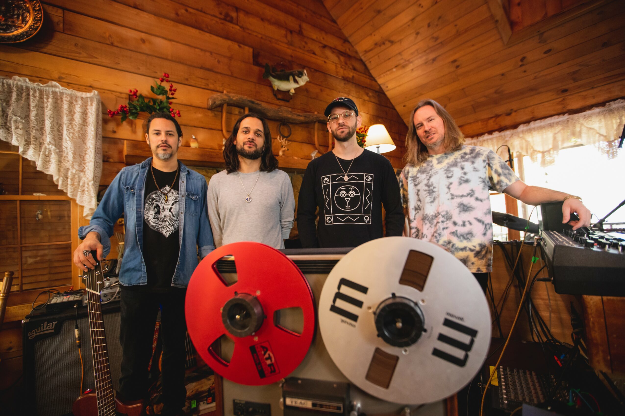 Four men stand in a wooden room with musical equipment, smiling at the camera.