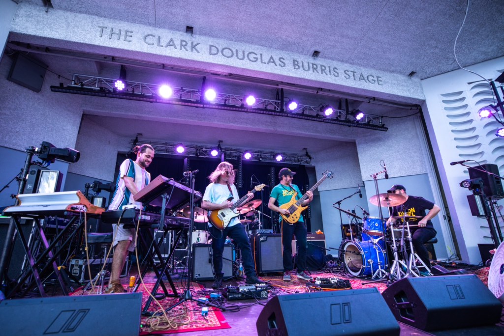 Band performs on stage with four musicians playing instruments, stage lights, and sound equipment, in a venue named The Clark Douglas Burris Stage.