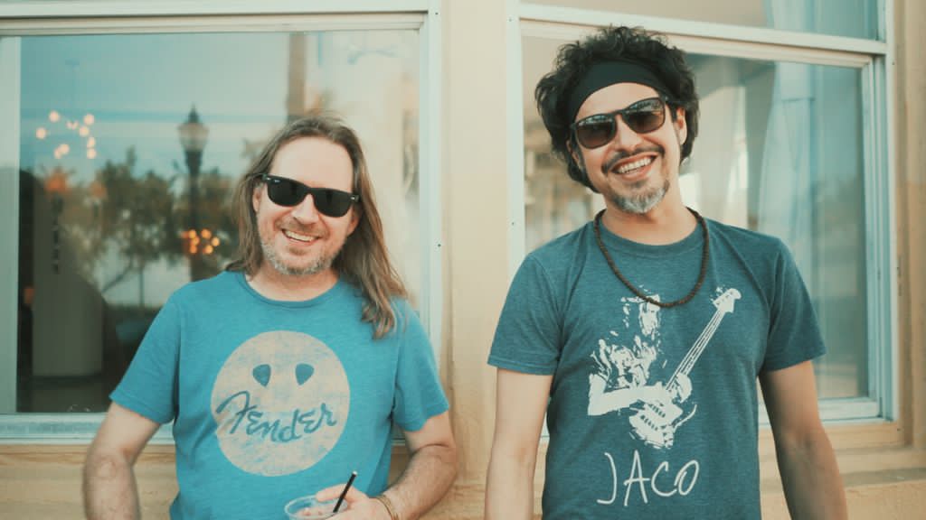 Two smiling men wearing sunglasses stand outside a building with large windows, one with long hair and a Fender shirt, the other with curly hair and a guitar shirt.