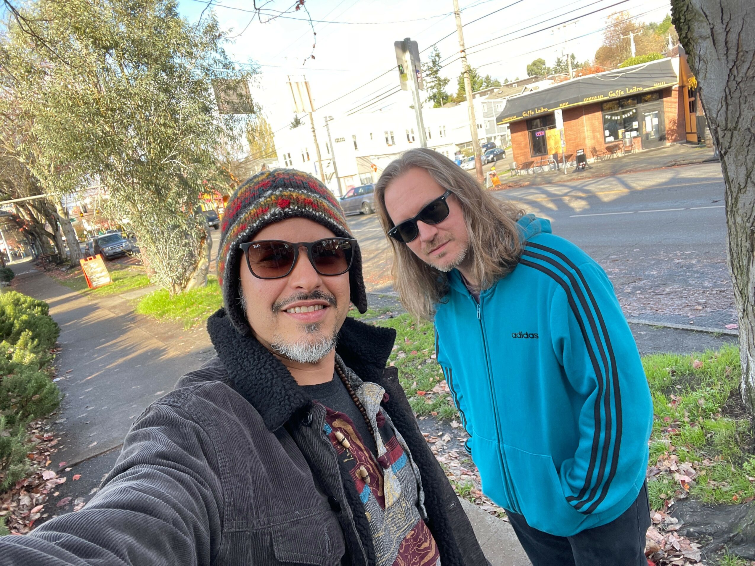 Two women smiling outdoors, wearing sunglasses and winter clothing, with trees and a street in the background.