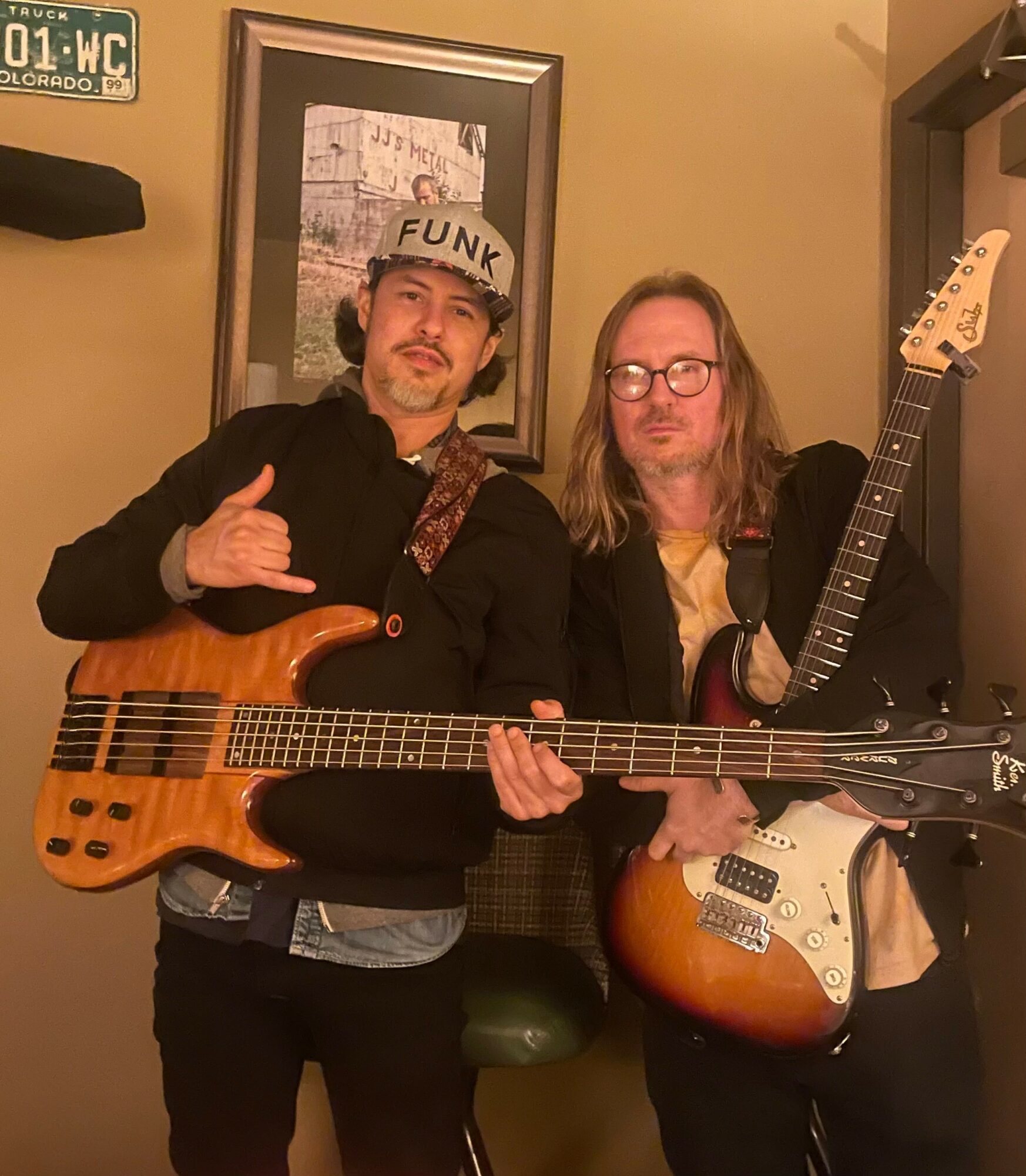 Two men holding guitars in a room with a framed picture on the wall, one wearing a cap with 'FUNK' and making a hand gesture.