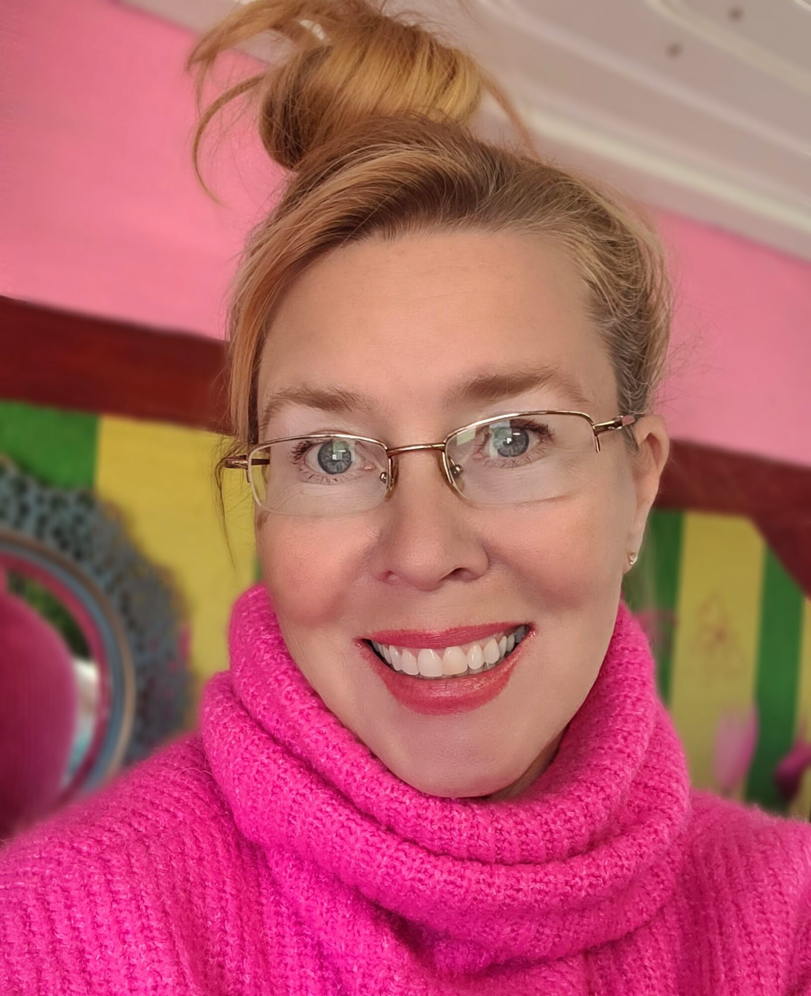 Woman with glasses smiling, wearing a pink turtleneck, in a colorful room with pink and green walls.