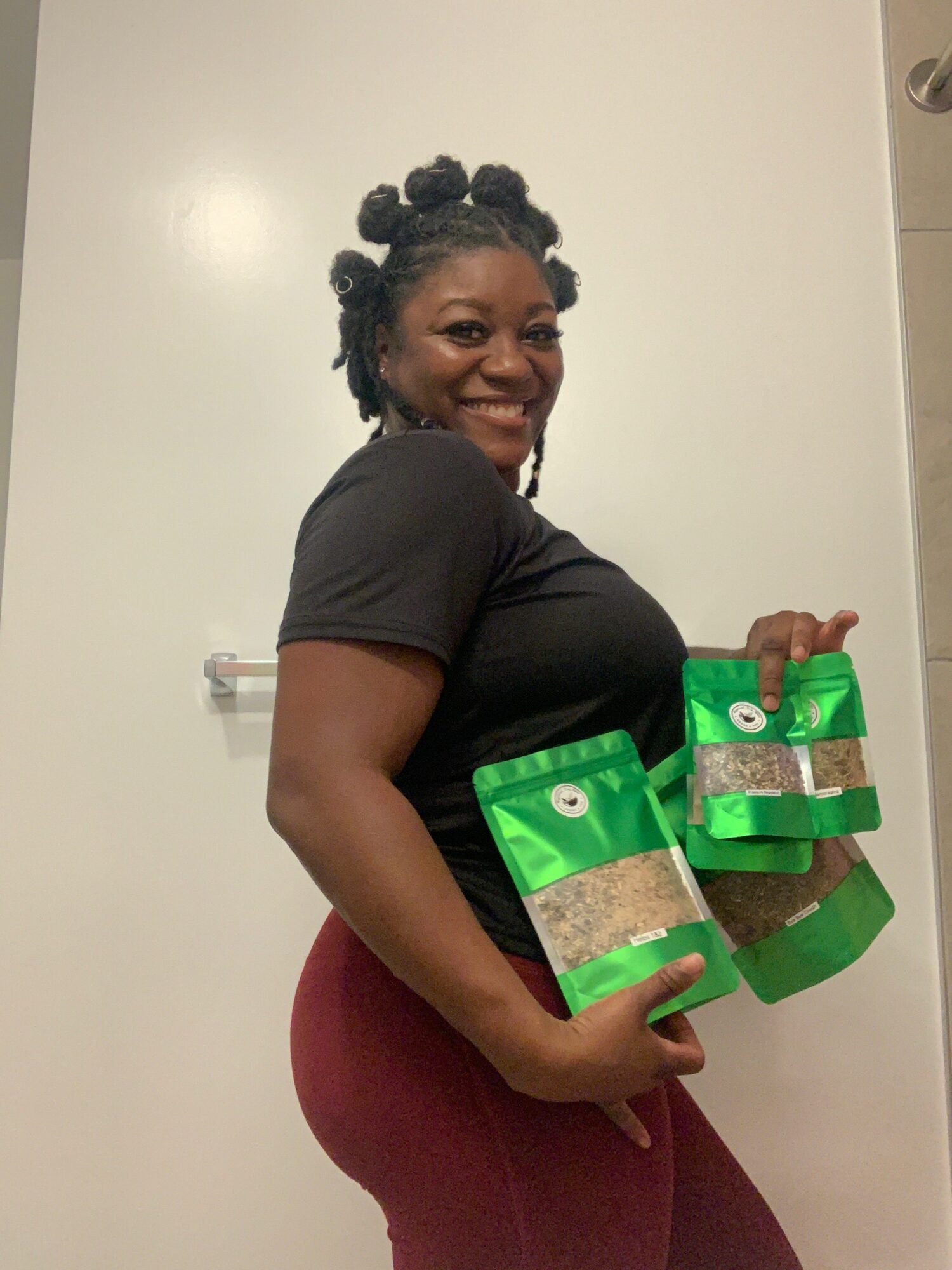 Woman holding multiple green snack bags, standing against a plain white wall, smiling, with dark hair styled in twists.
