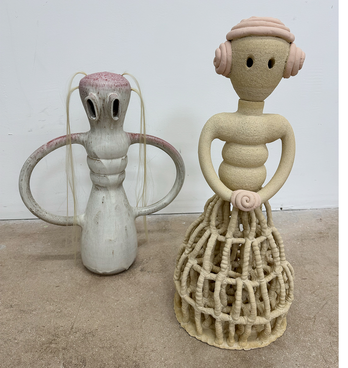 Two handmade figurines, one with a tall, rounded body and large arms, the other with a head, torso, and a woven skirt, standing on a floor against a white wall.