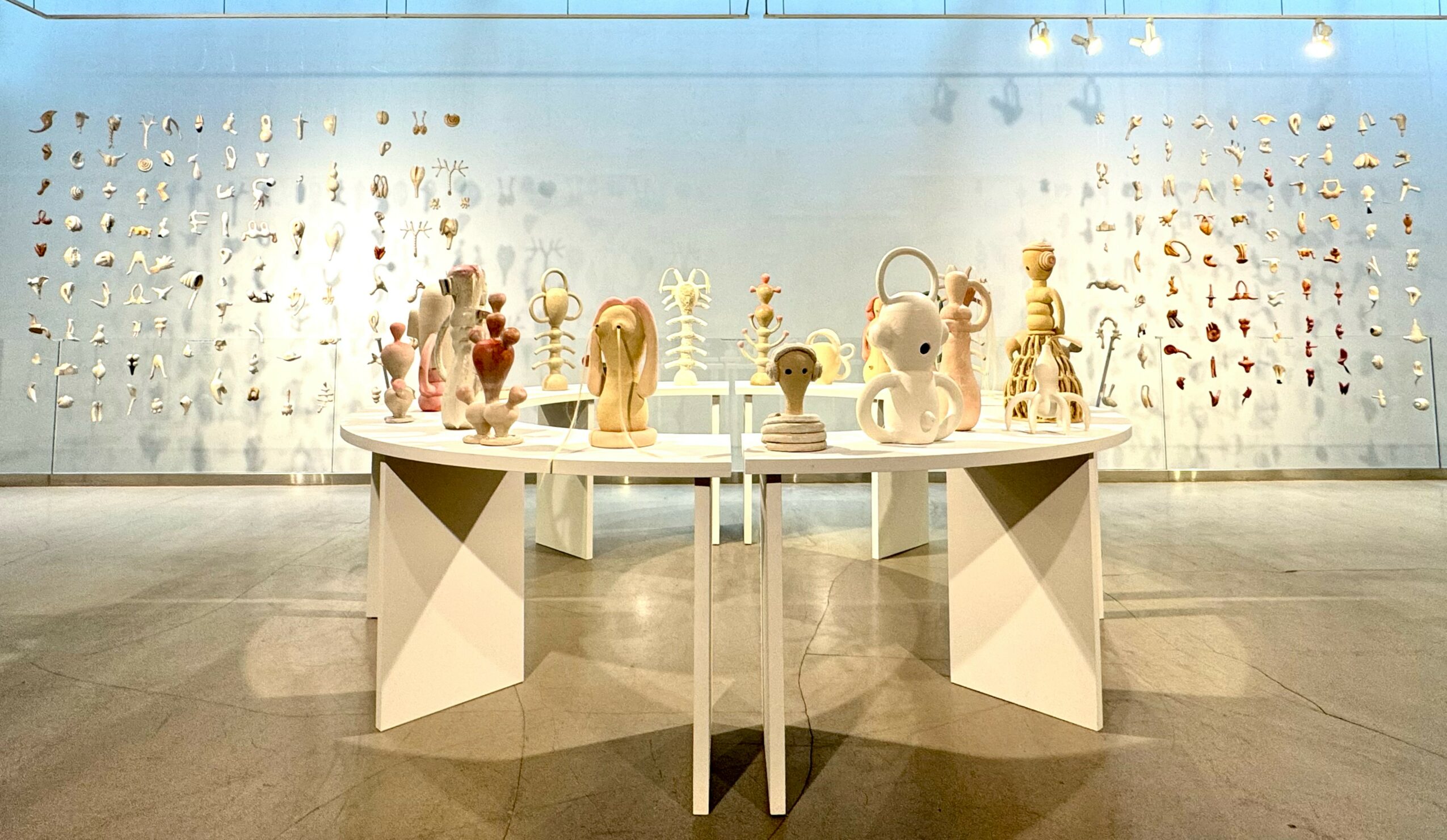 Display of ceramic sculptures on two white tables in front of a wall with small objects, in a gallery setting.