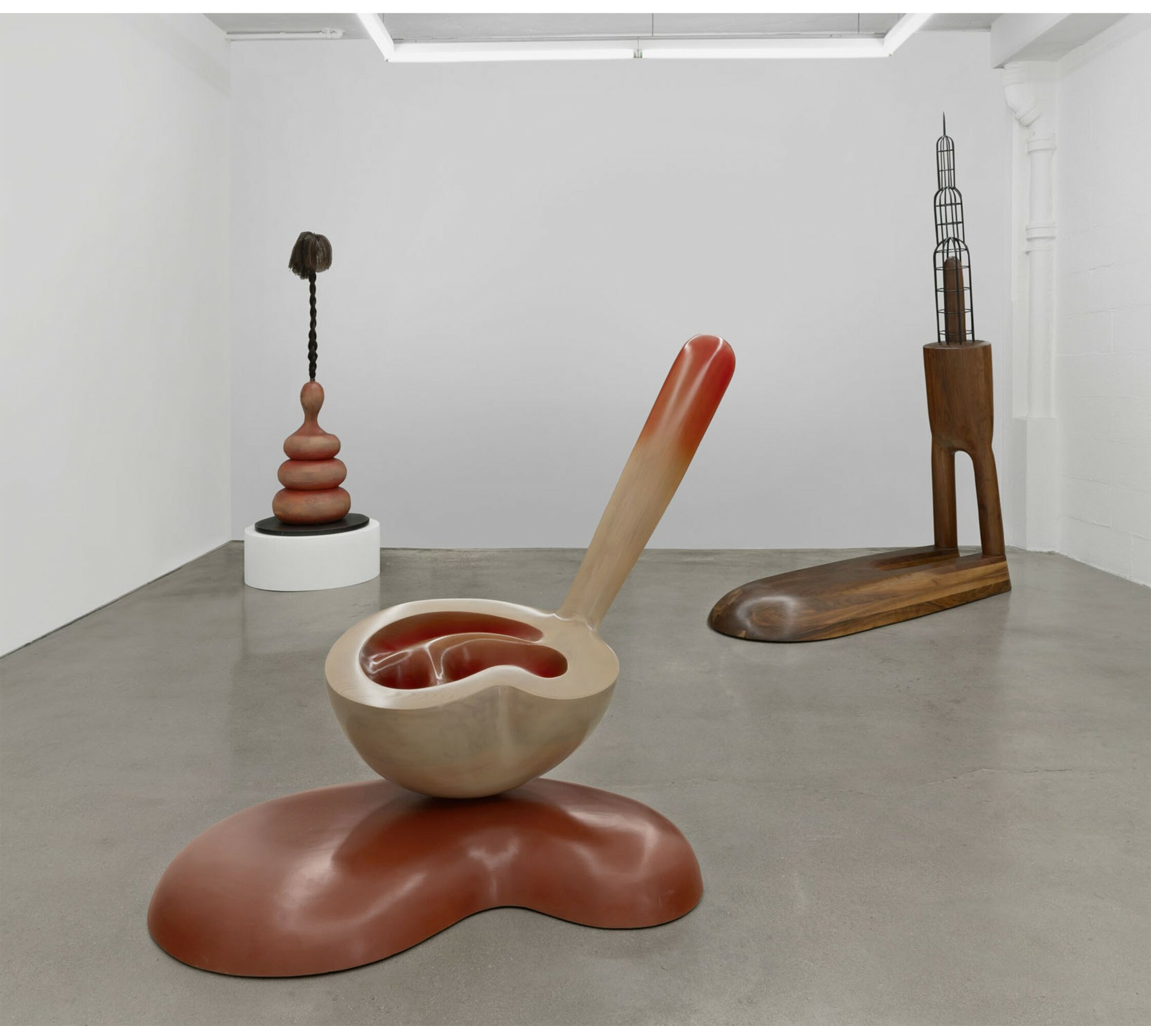 Three abstract sculptures in a gallery with white walls and concrete floor, one with a rounded base and elongated handle, others with geometric shapes.