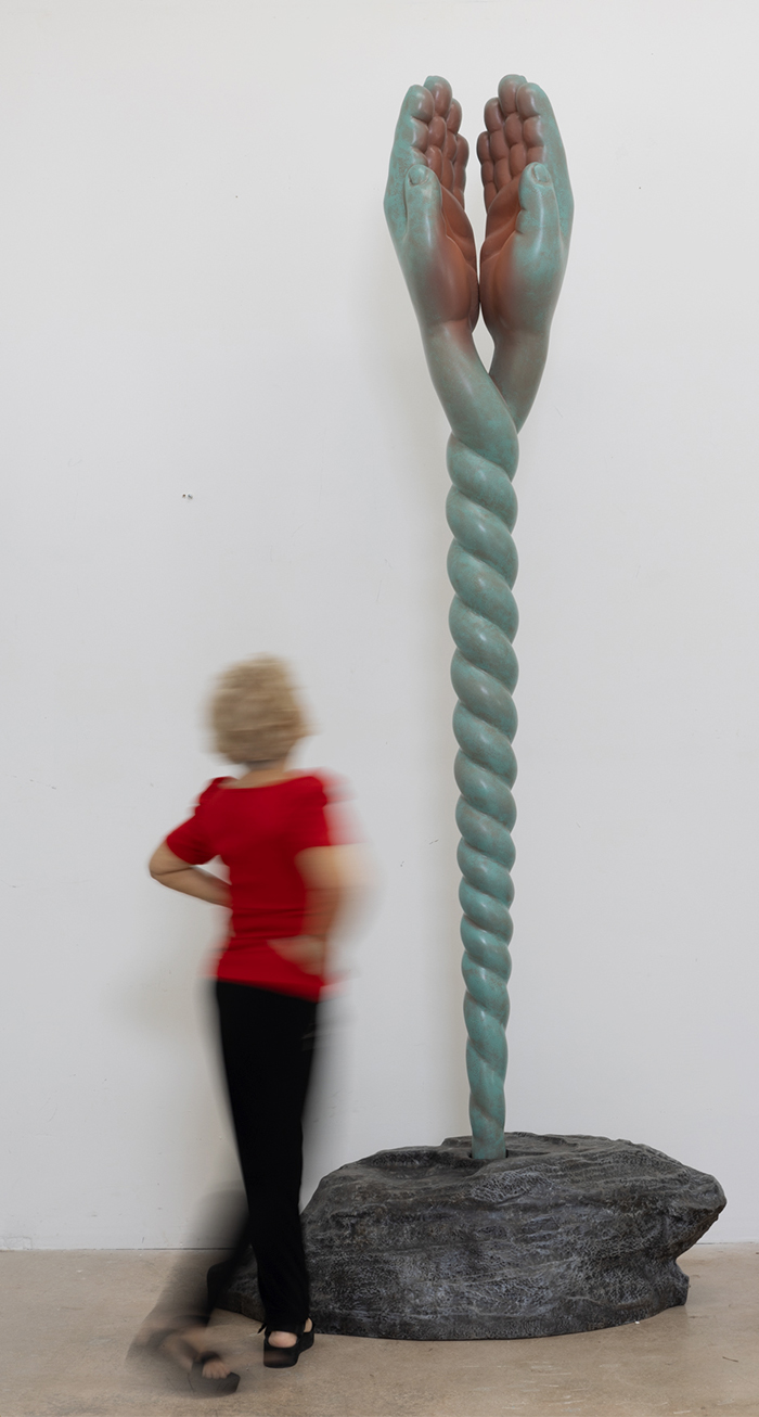Sculpture of intertwined hands holding a twisted column, mounted on a rock base, with a person in a red shirt walking nearby.