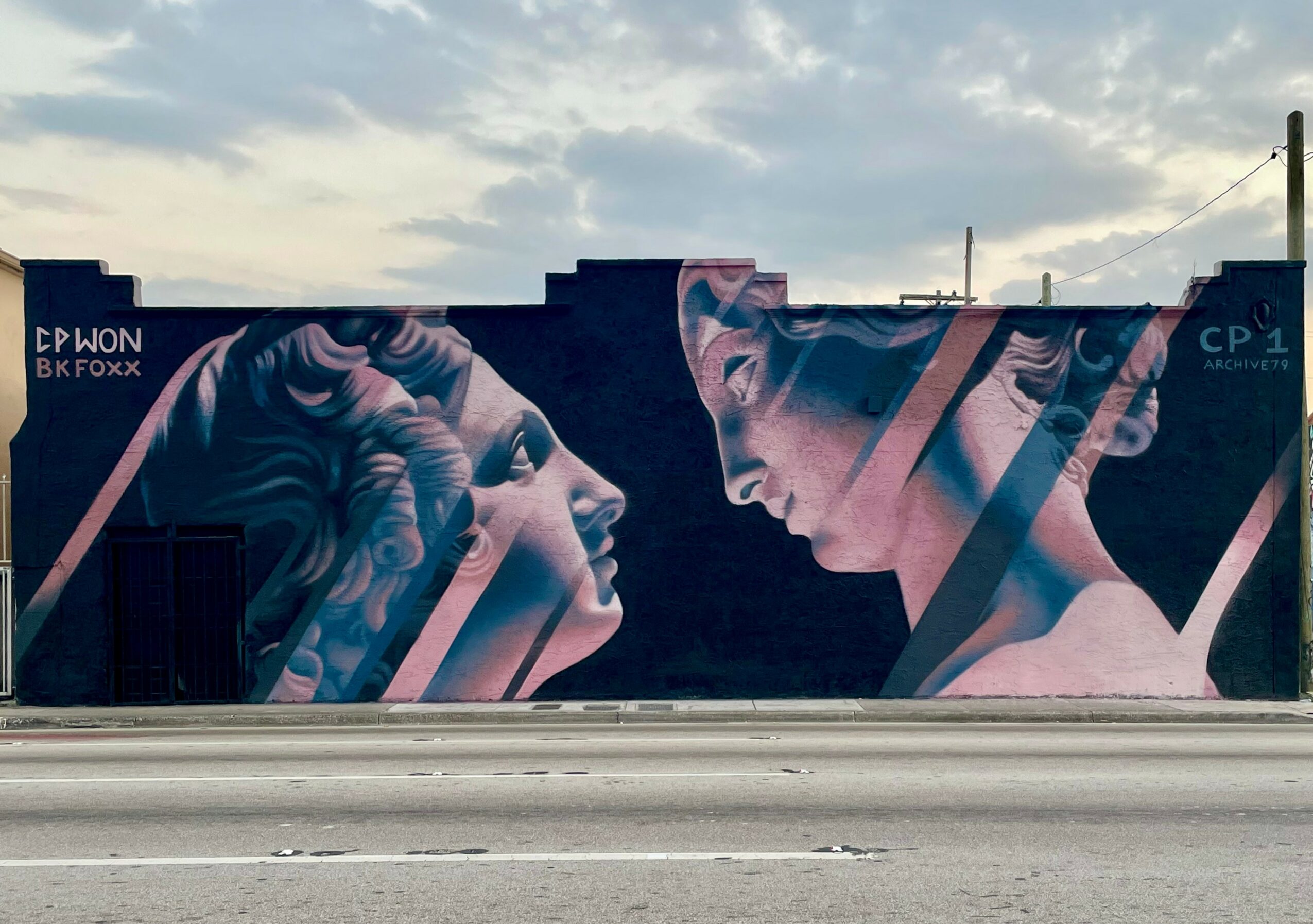 Street mural depicting two faces, one looking upward and the other downward, with a cloudy sky background.