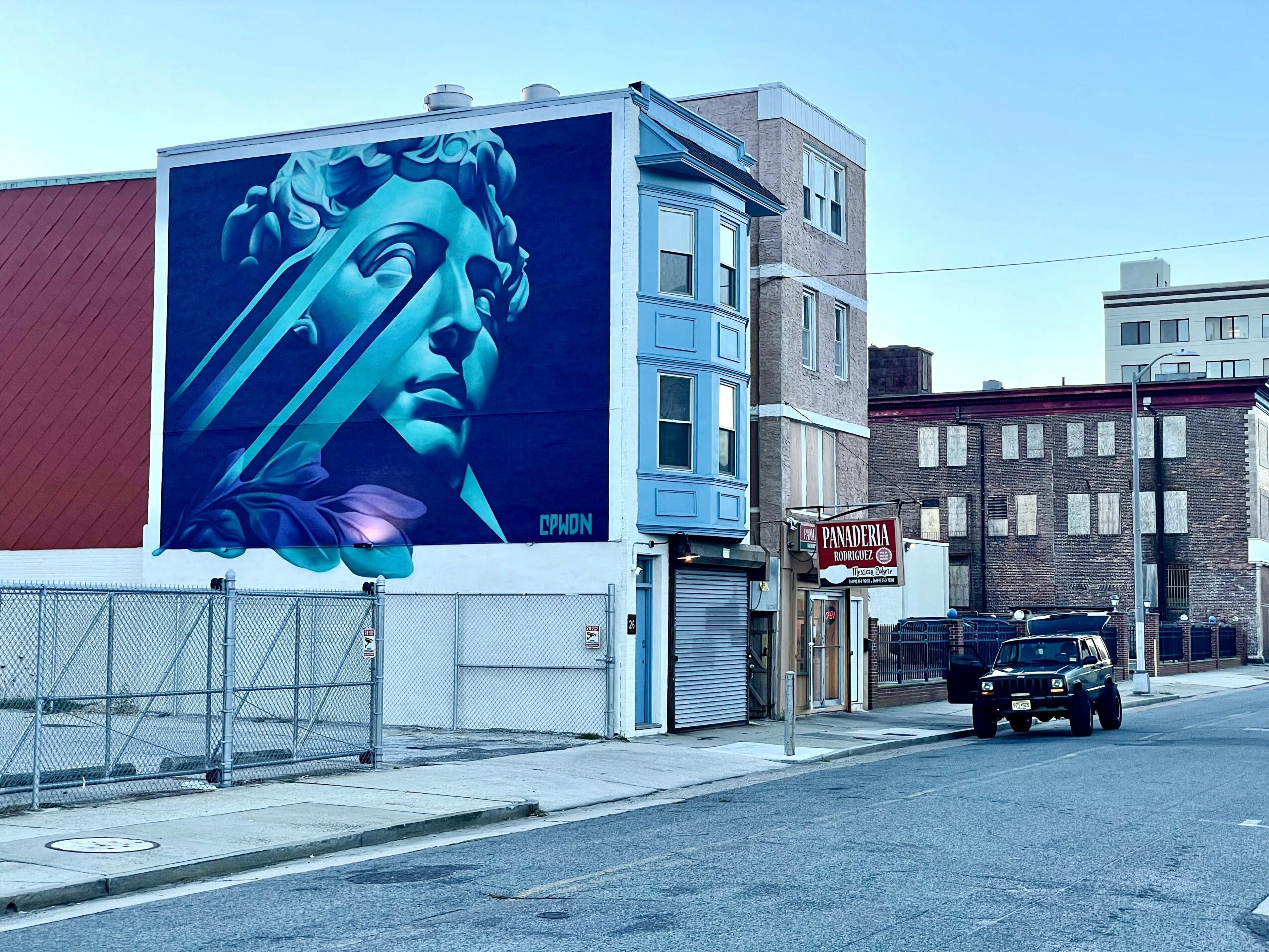 Street scene with a large blue mural of a woman's face on a building, parked car, and surrounding buildings.