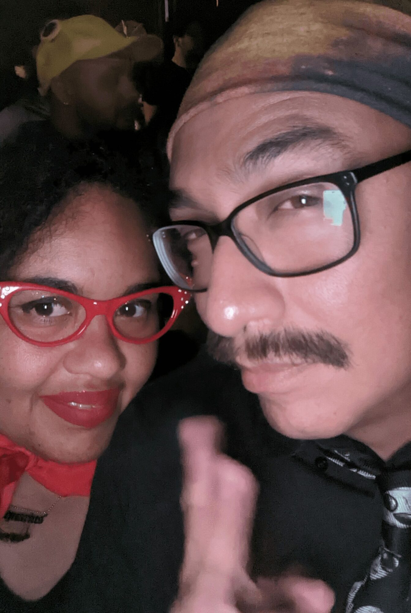 Two people close together, one with red glasses and the other with glasses and a head covering, in a dark setting.