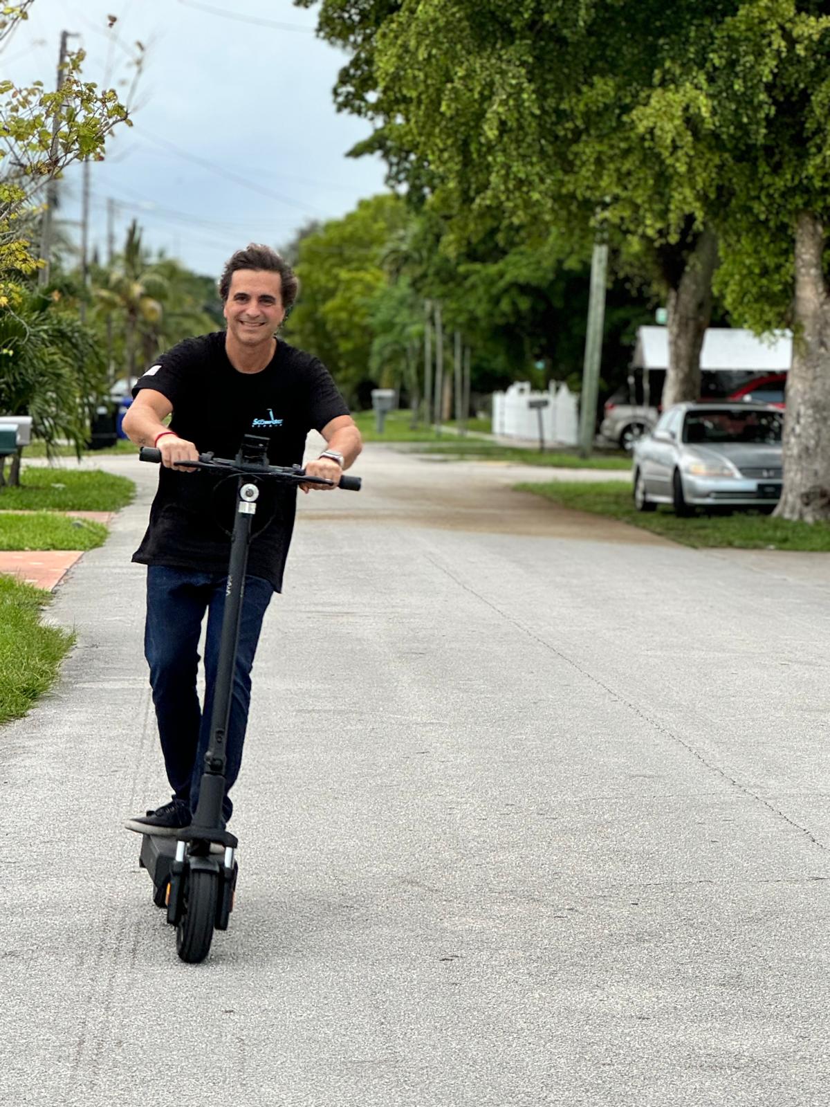 Person riding an electric scooter on a residential street, smiling, with trees and parked cars in the background.
