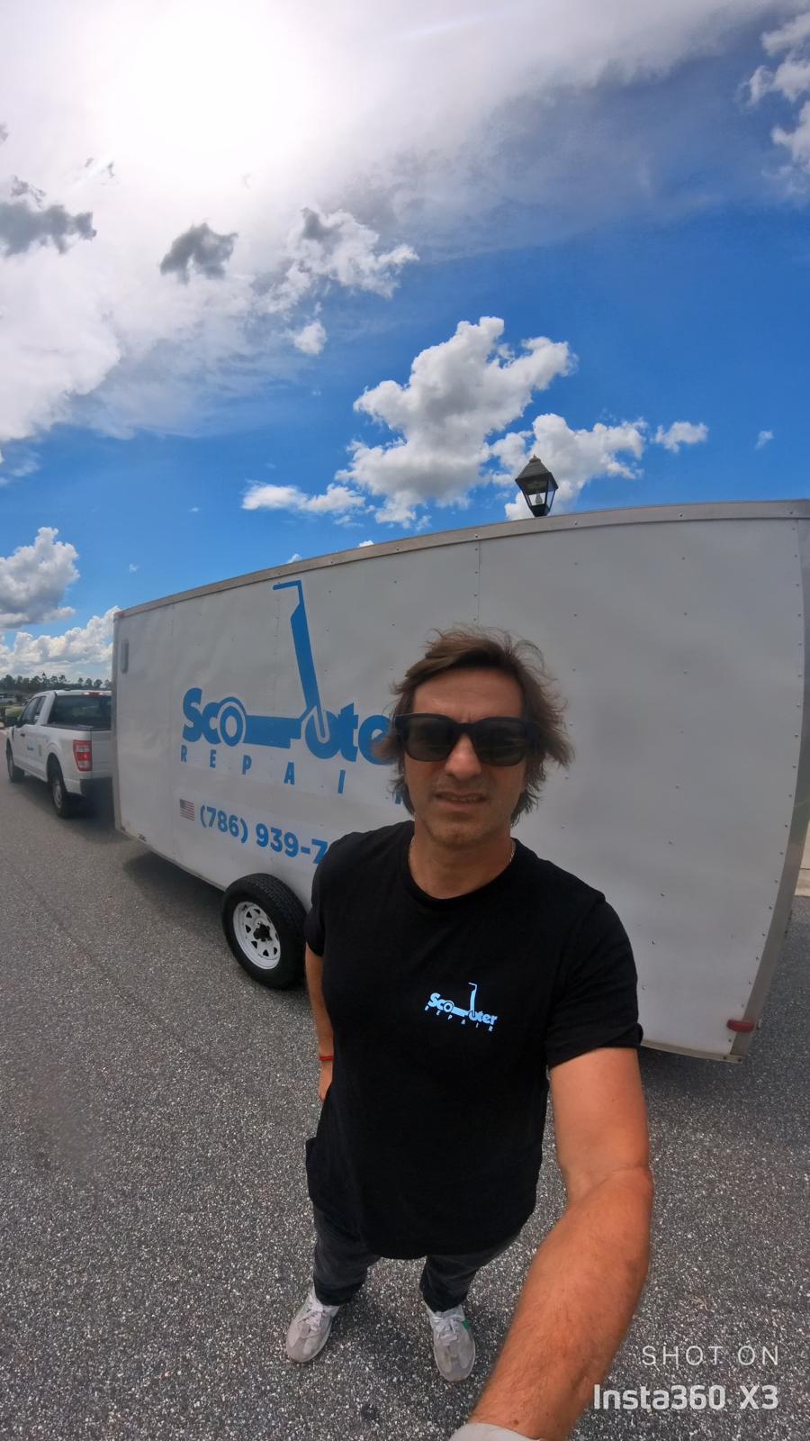 Person with sunglasses standing outdoors near a trailer and parked cars under a partly cloudy sky.