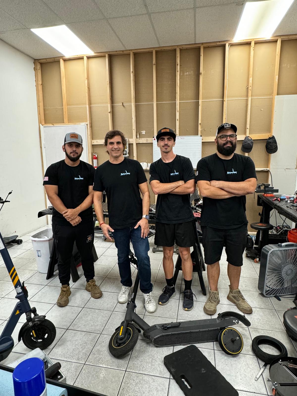 Four people stand in a room with a partially finished wall, surrounded by equipment and a scooter, smiling at the camera.
