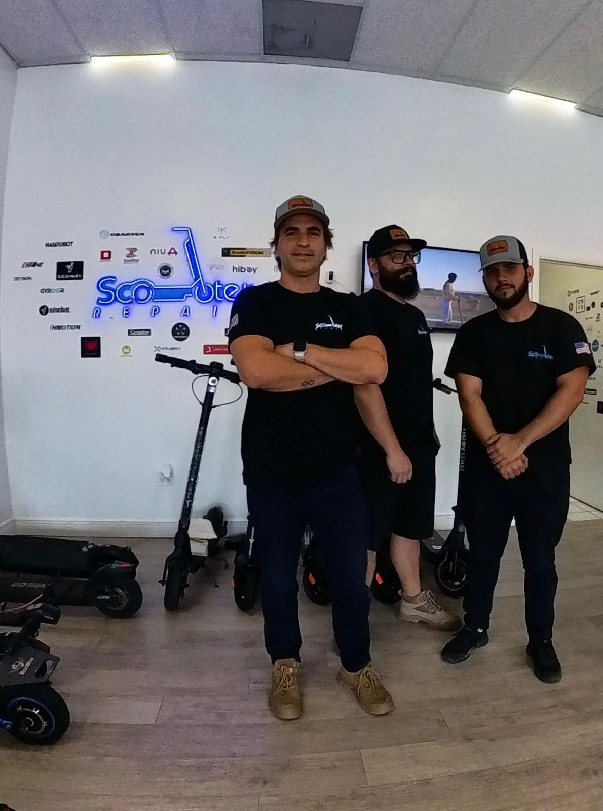 Three men standing indoors in front of a wall with a logo, with scooters on the floor, facing forward.