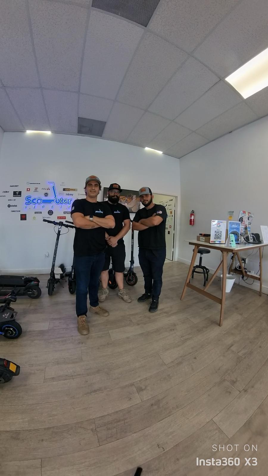 Four people standing in a room with a whiteboard, table, and equipment, facing the camera with arms crossed.
