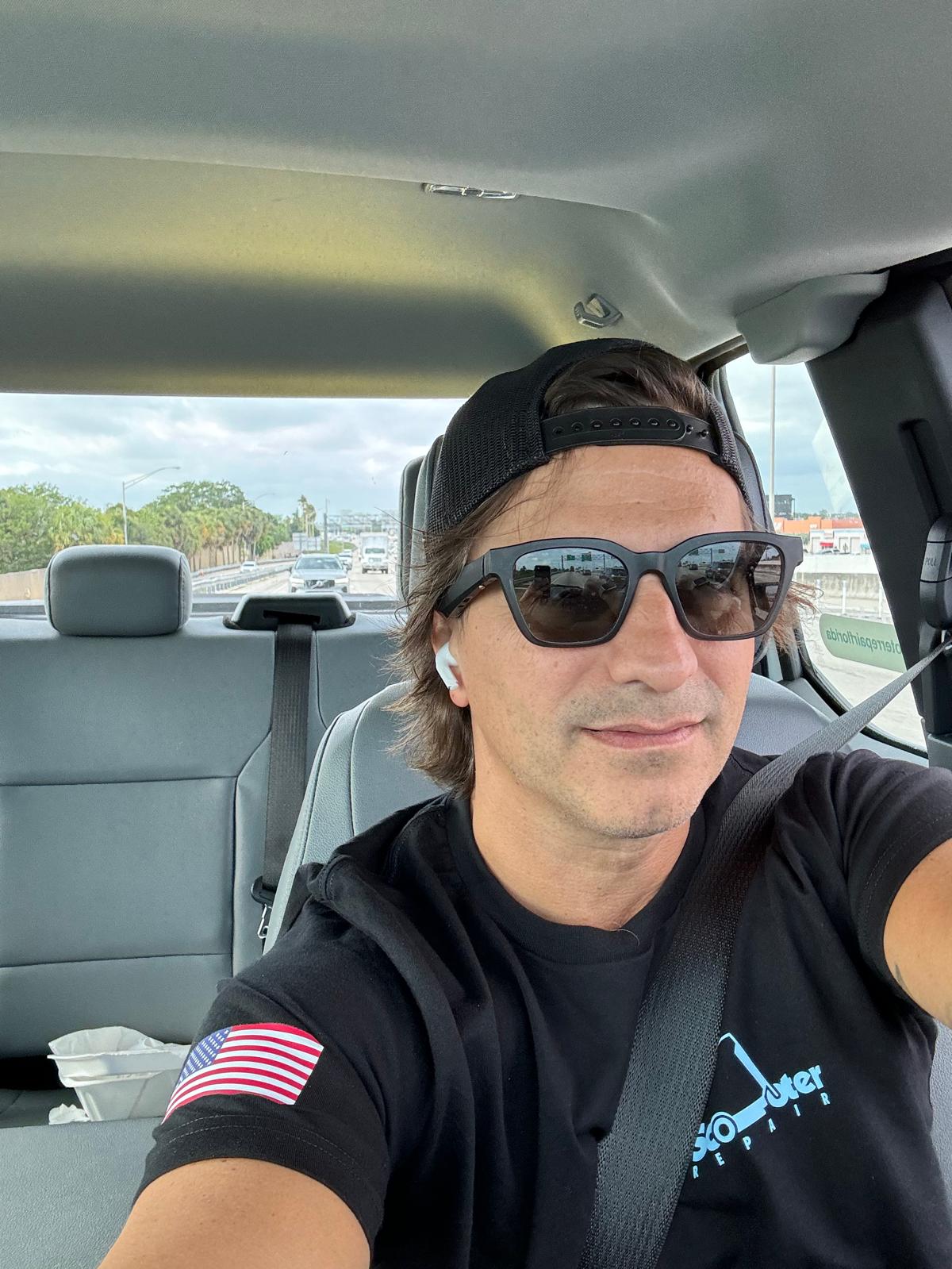 Man wearing sunglasses and a black cap, sitting in a car, with a highway view outside.