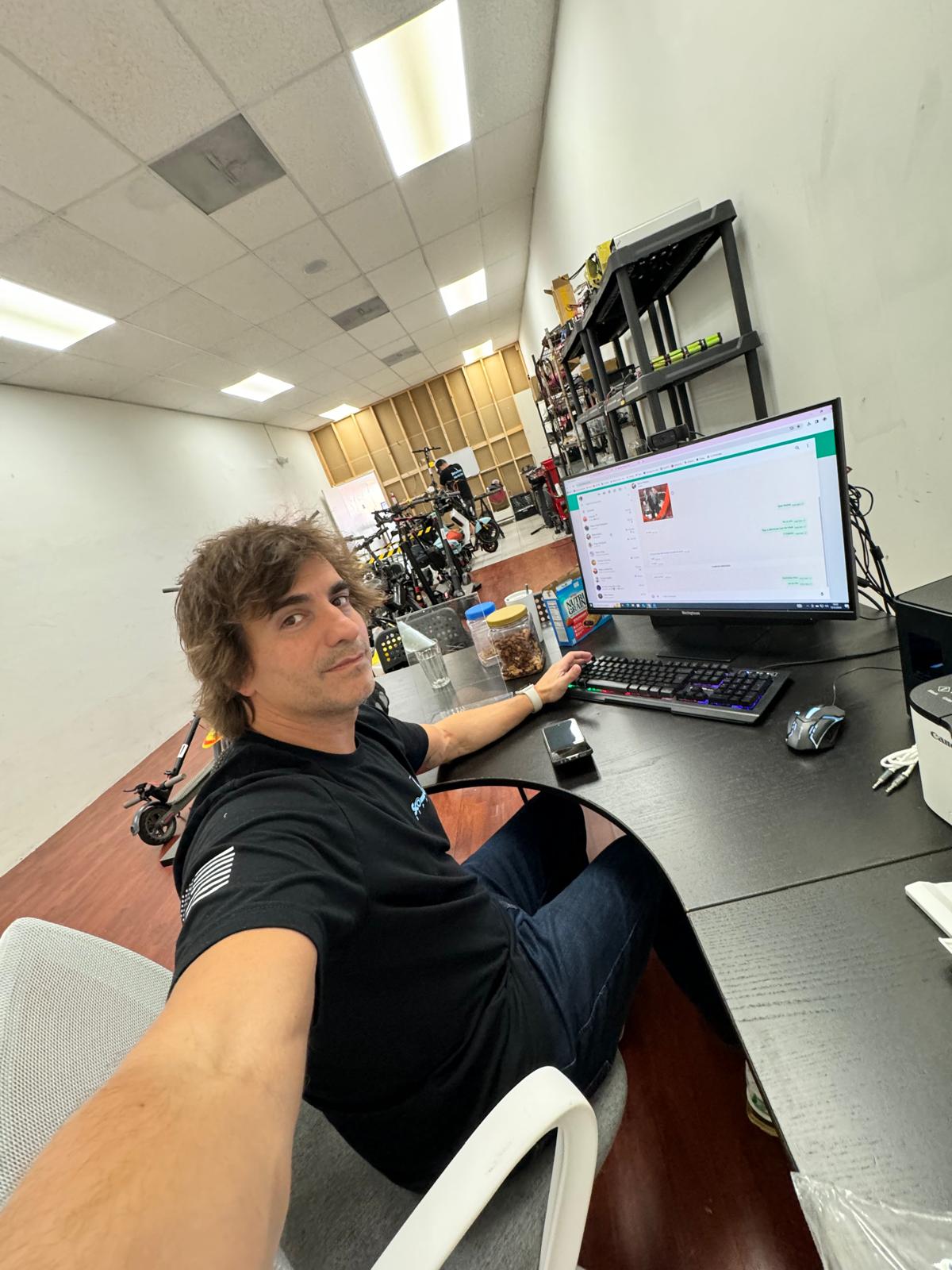 Young man with brown hair sitting at a desk in an office, taking a selfie with a computer monitor and office supplies visible.