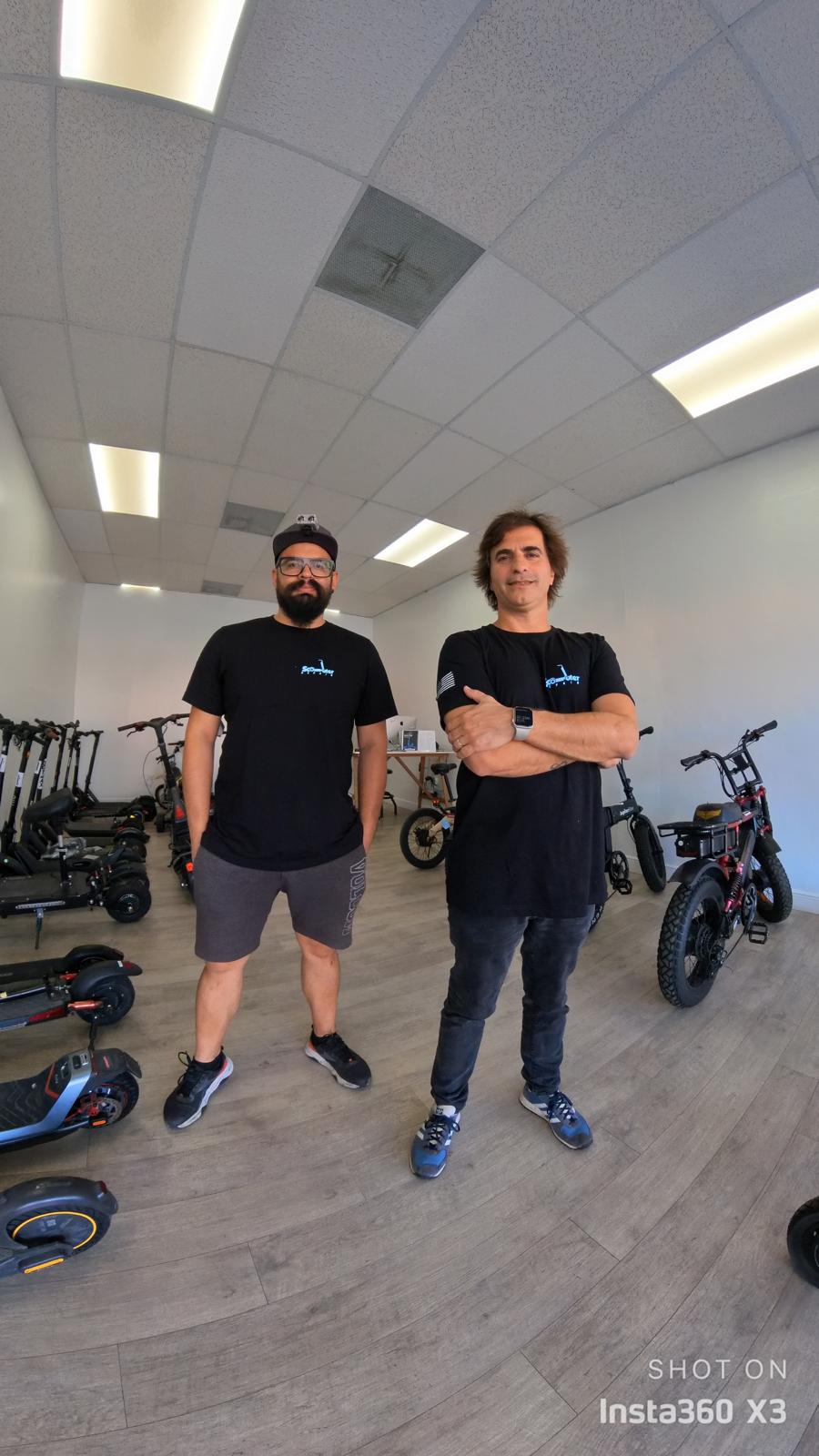Two men stand in a room with bicycles and scooters, ceiling with lights, one with arms crossed, the other with hands in pockets.
