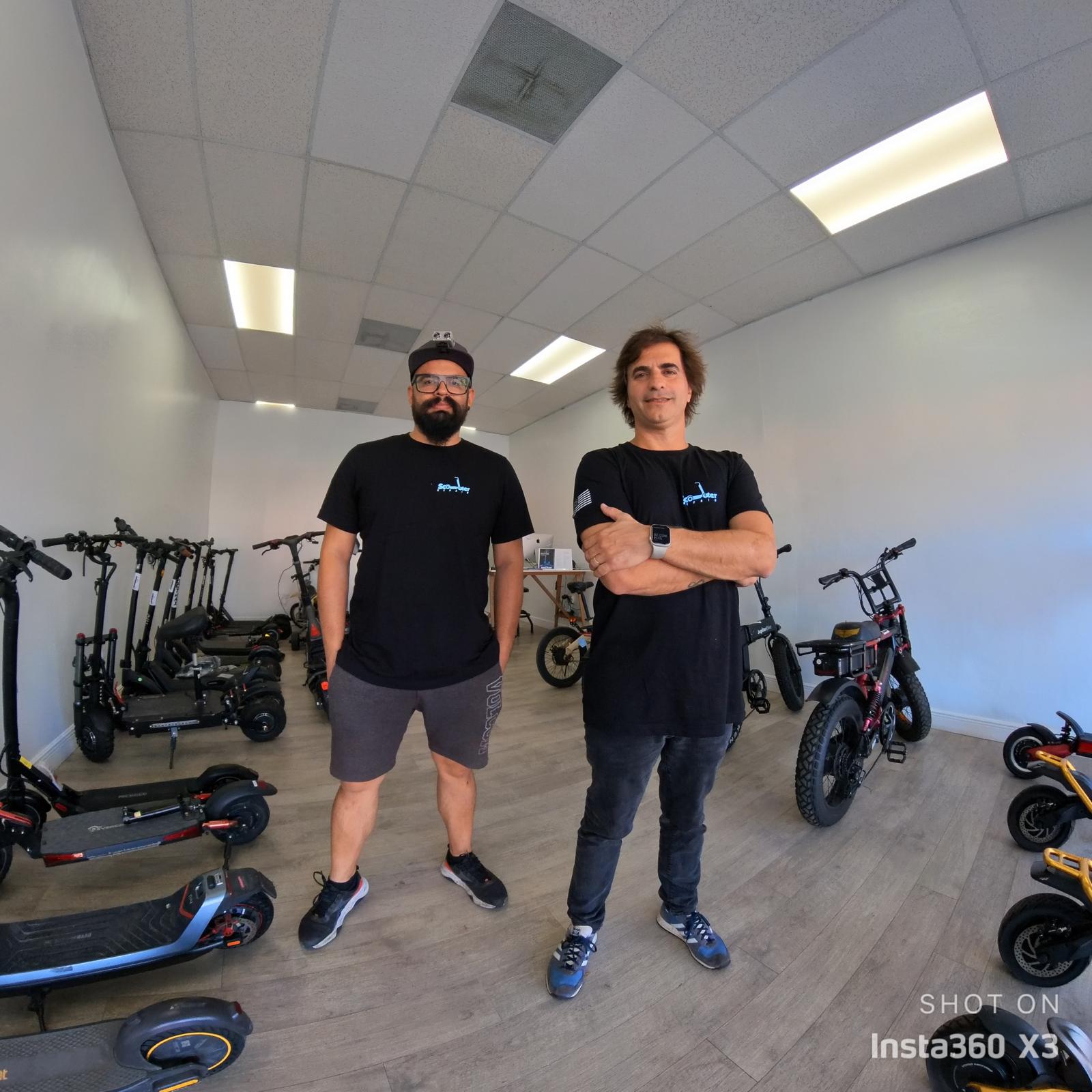Two men stand in a room with electric scooters and bikes, white walls, and ceiling lights, facing the camera.