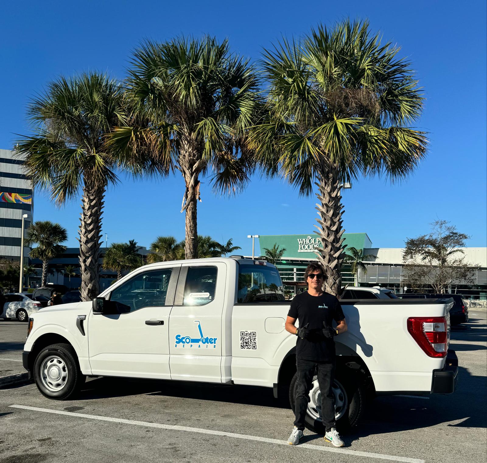Person standing next to a white pickup truck with palm trees and a building in the background, sunny day.