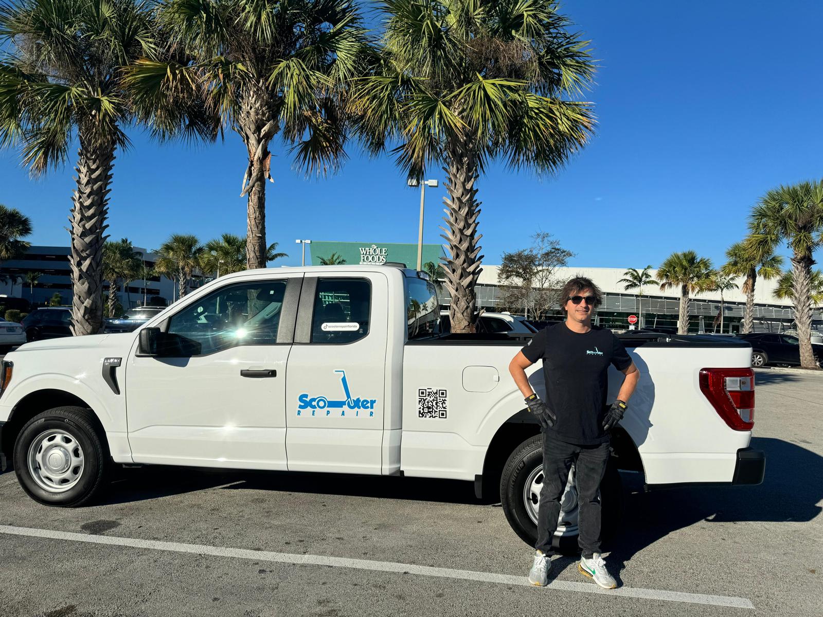 Person standing next to a white pickup truck with trees and a parking lot in the background.