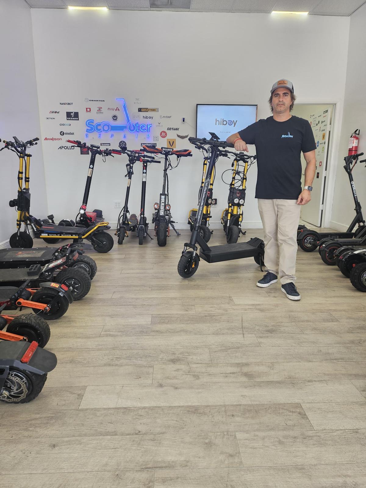 Person standing in a room with electric scooters and a display screen, white walls, wooden floor, and bright lighting.
