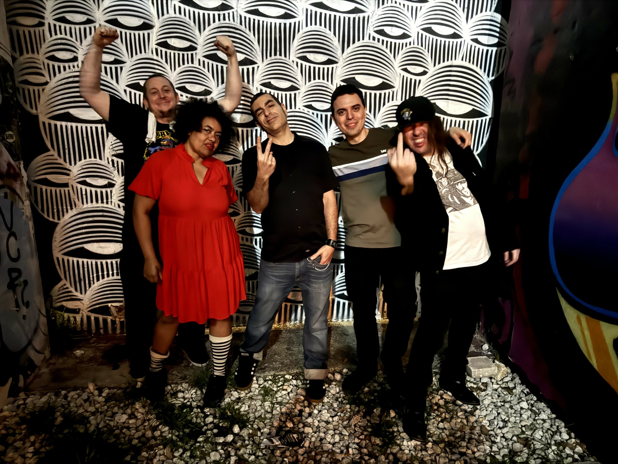 Group of five people standing outdoors in front of a patterned wall, smiling and making gestures, with gravel ground.