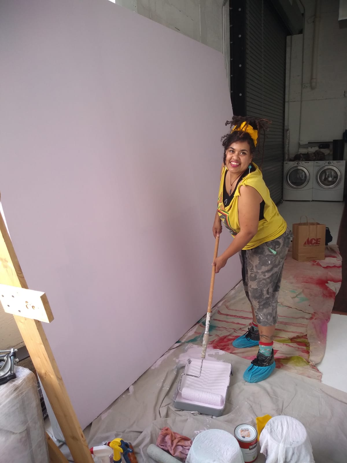Young girl smiling, holding a mop, standing on a plastic sheet in front of a large white wall, wearing a yellow shirt and patterned pants.