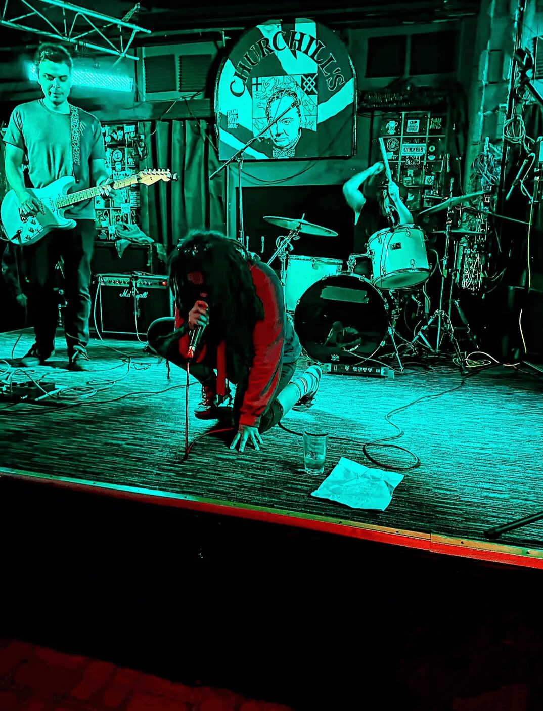 Person kneeling on stage with microphone, band playing instruments in background, stage with sign and equipment, dim lighting.