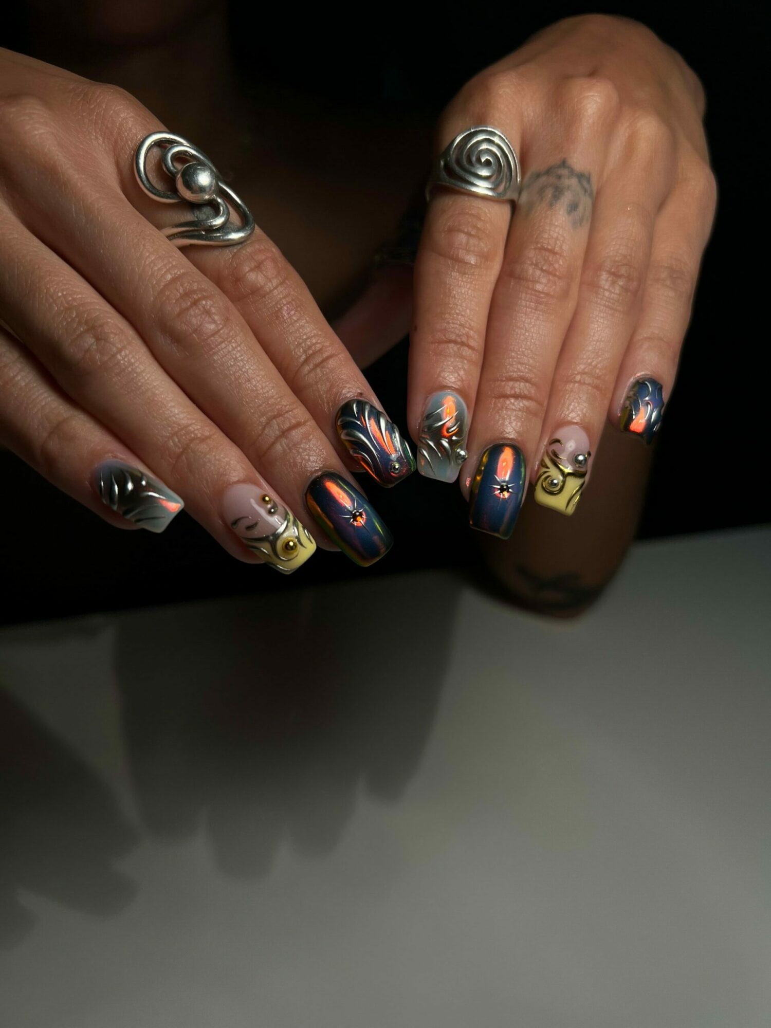 Two hands with colorful, patterned nail polish and large silver rings, against a dark background.