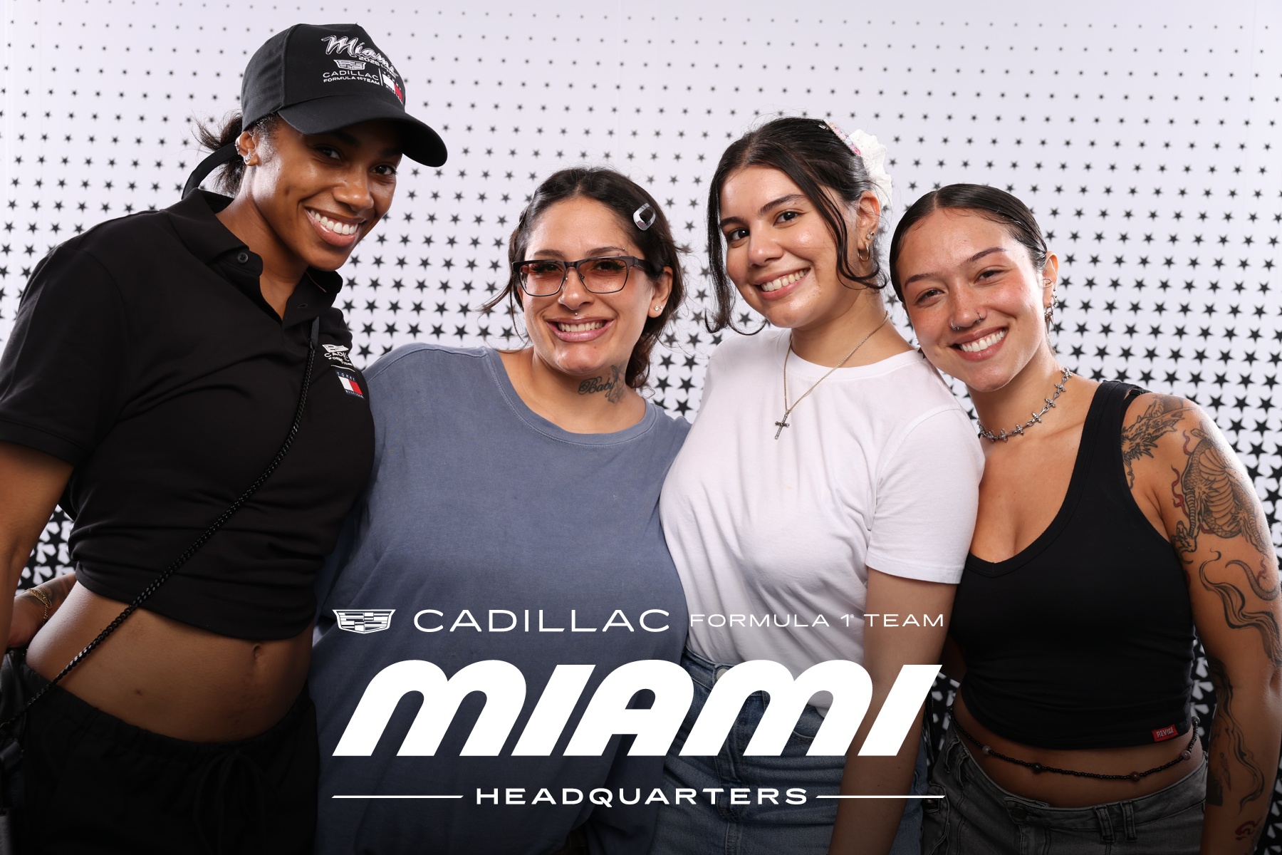 Four women smiling, standing together in front of a white perforated wall, wearing casual athletic clothing.