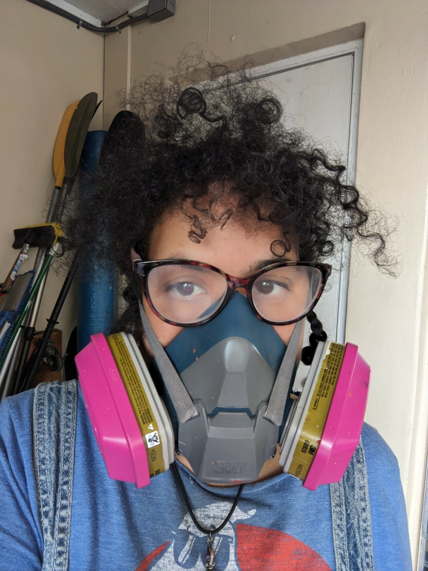 Person with curly hair wearing glasses and a respirator mask with pink and yellow filters.