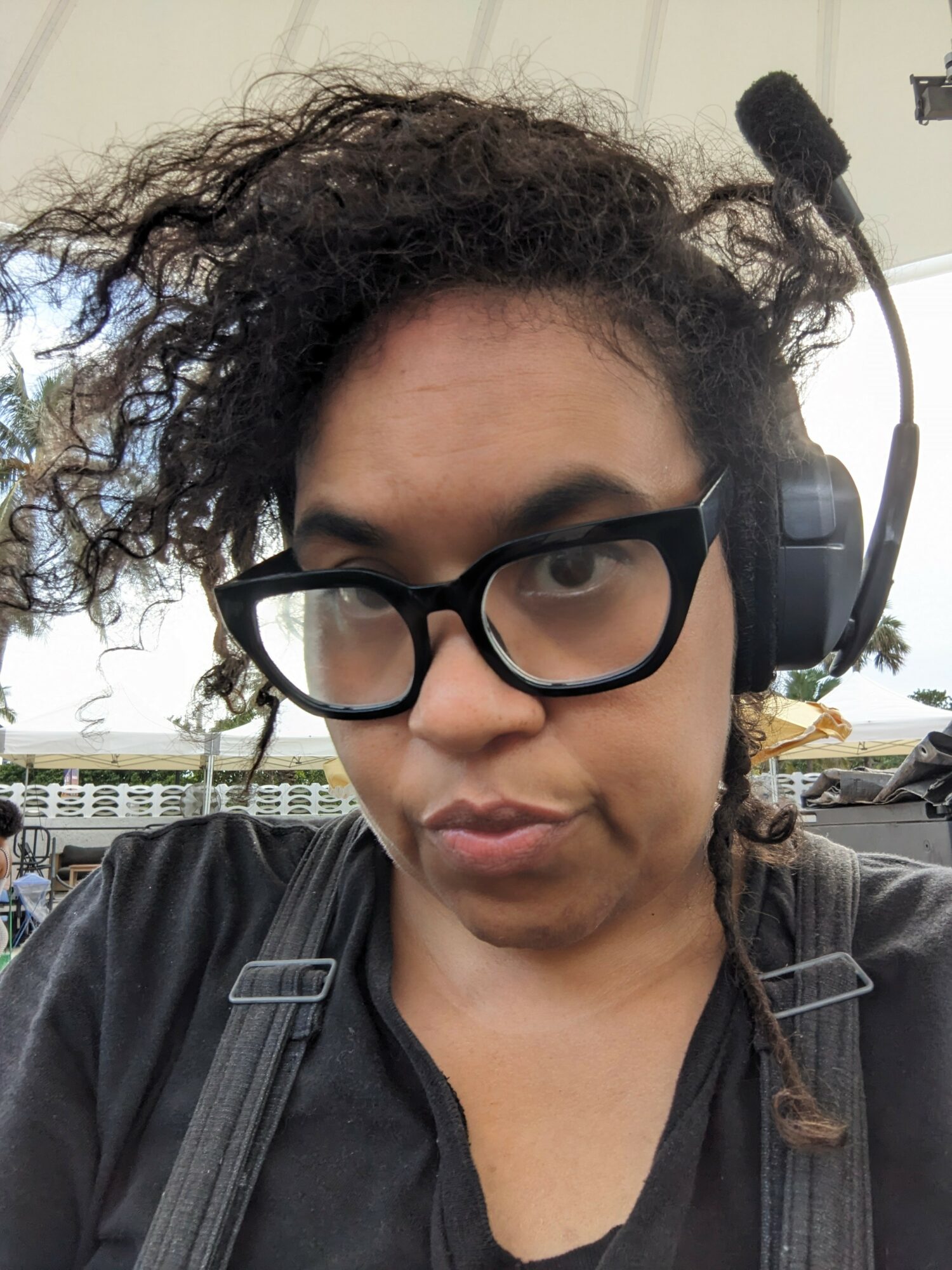 Person with curly hair, glasses, and headset outdoors, looking at camera.