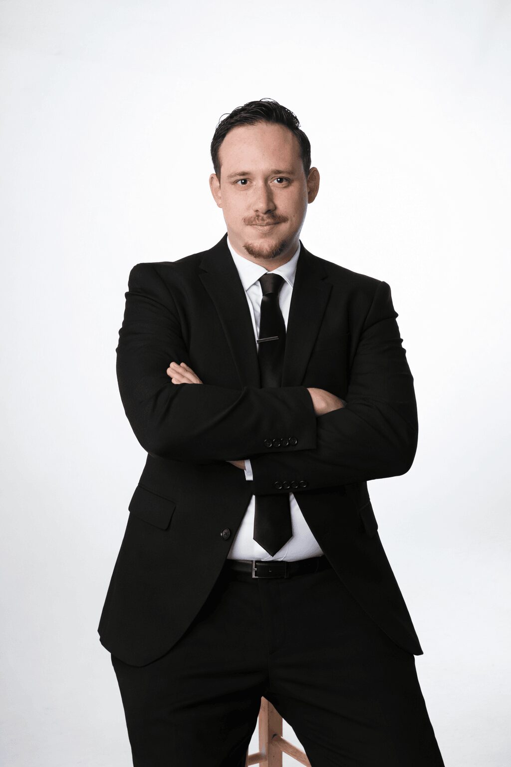 Man in black suit with arms crossed, standing against a white background.