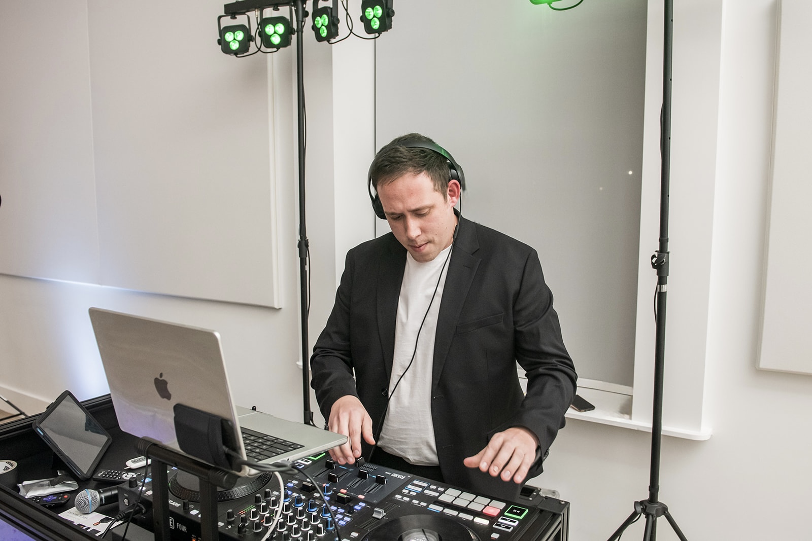 A man in a black blazer and white shirt DJing with equipment, laptop, and headphones in a room with lighting equipment.