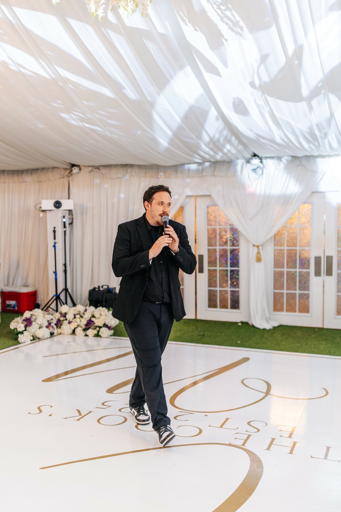 Man in black suit speaking into microphone on decorated event stage with floral arrangements and white curtains.