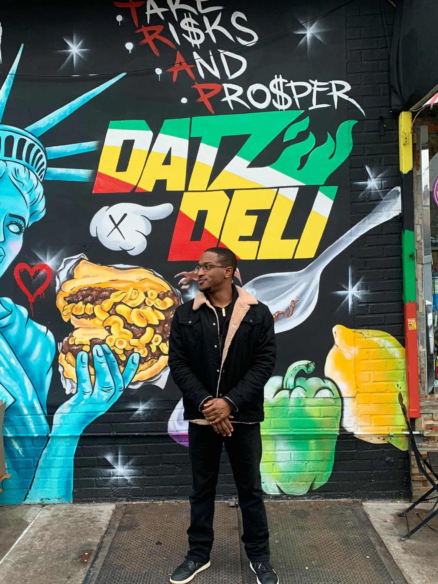 Man standing in front of colorful graffiti mural with Statue of Liberty, food, and text, outdoors on a city street.