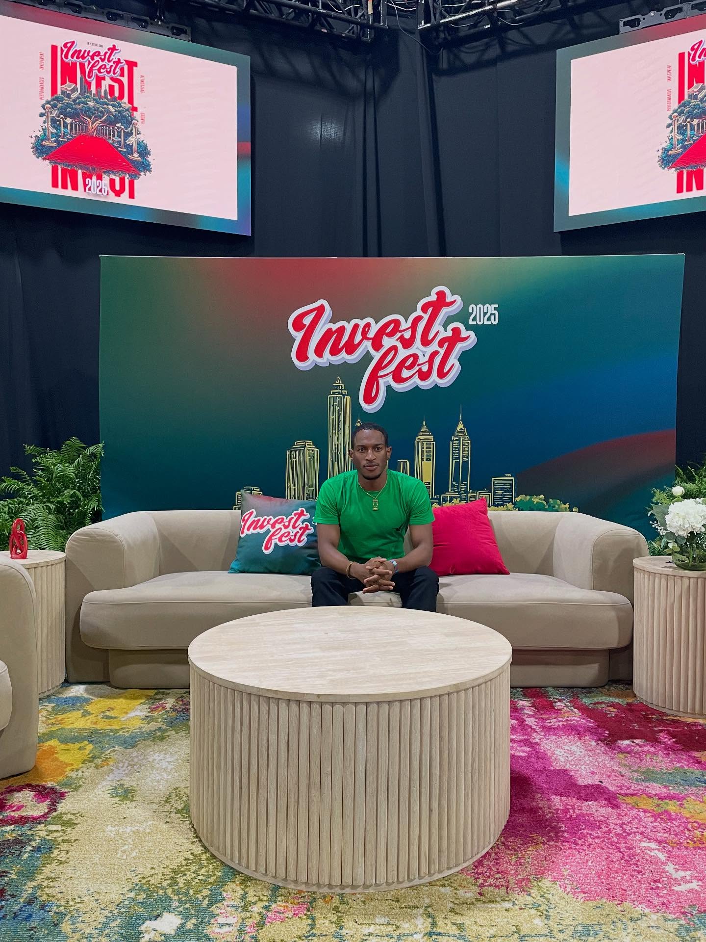 Person sitting on a beige sofa with pink and white pillows, in front of a colorful backdrop with 'Invest Fest' logo, two screens above, and a round table in front.